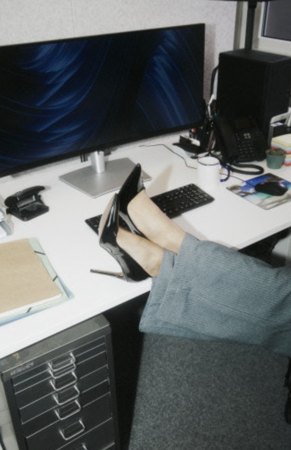 Person with legs crossed wearing high heels and gray pants at a cluttered office desk with a computer, monitor, keyboard, and office supplies.