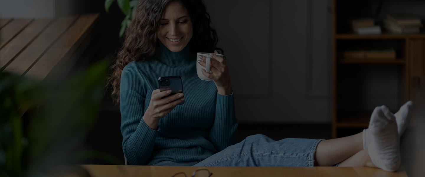 A woman sitting comfortably, smiling while looking at her phone and holding a mug in a cozy indoor setting.
