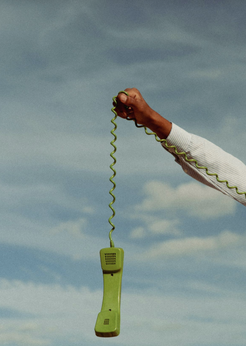 A hand holding a green corded telephone against a cloudy sky background.