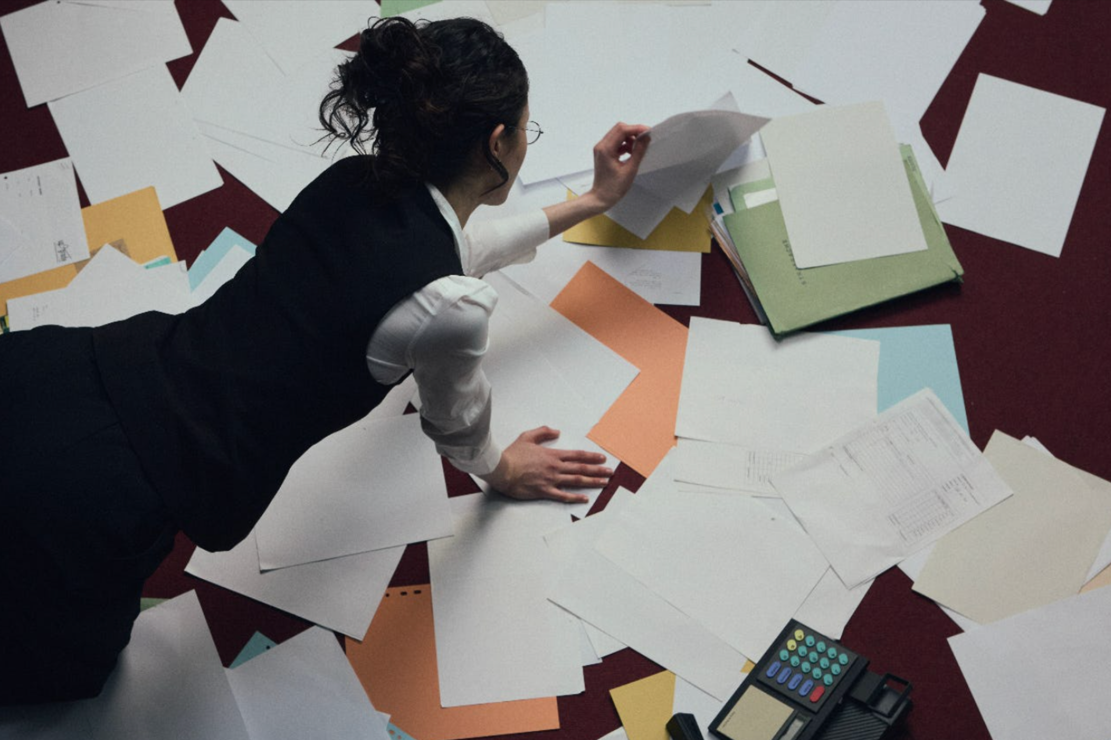 A woman in business attire lying on the floor amidst scattered papers, files, a calculator, and a folder.