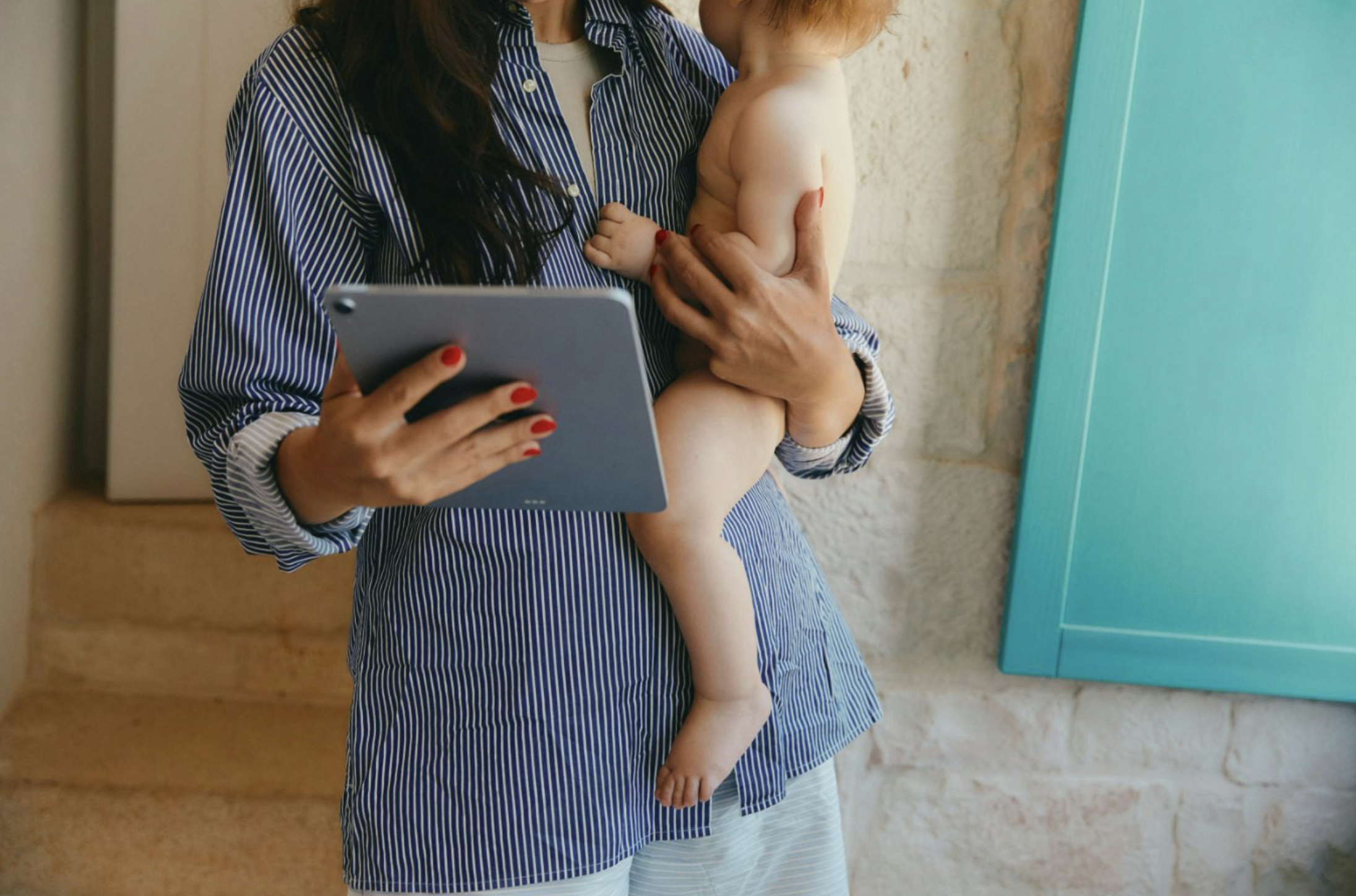 A woman holding a young child, with the child's back to the camera. The woman is reading or looking at a tablet, and the child is standing on the woman's hip. They are indoors, with a light-colored brick wall and a teal-colored object in the background.