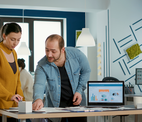 Two people working together at a modern office desk, reviewing documents and data on a laptop, with bright lighting and contemporary decor.