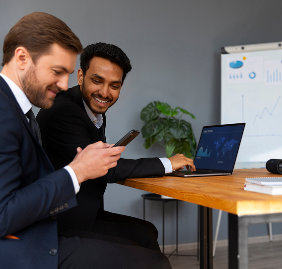 Two business professionals in suits looking at a smartphone in an office with a laptop and presentation charts in the background.