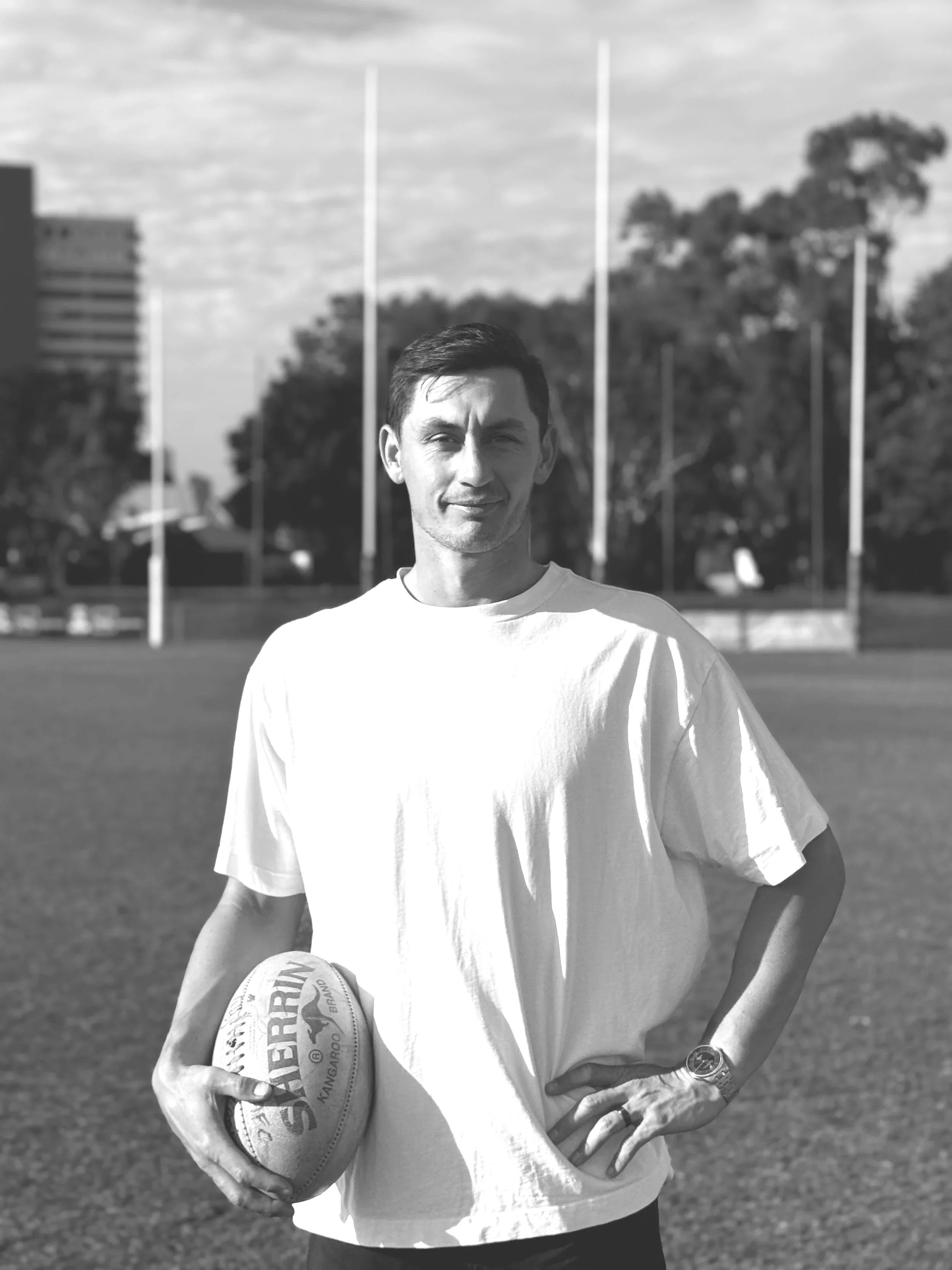 A young man standing on a sports field holding a rugby ball, with goalposts and trees in the background.