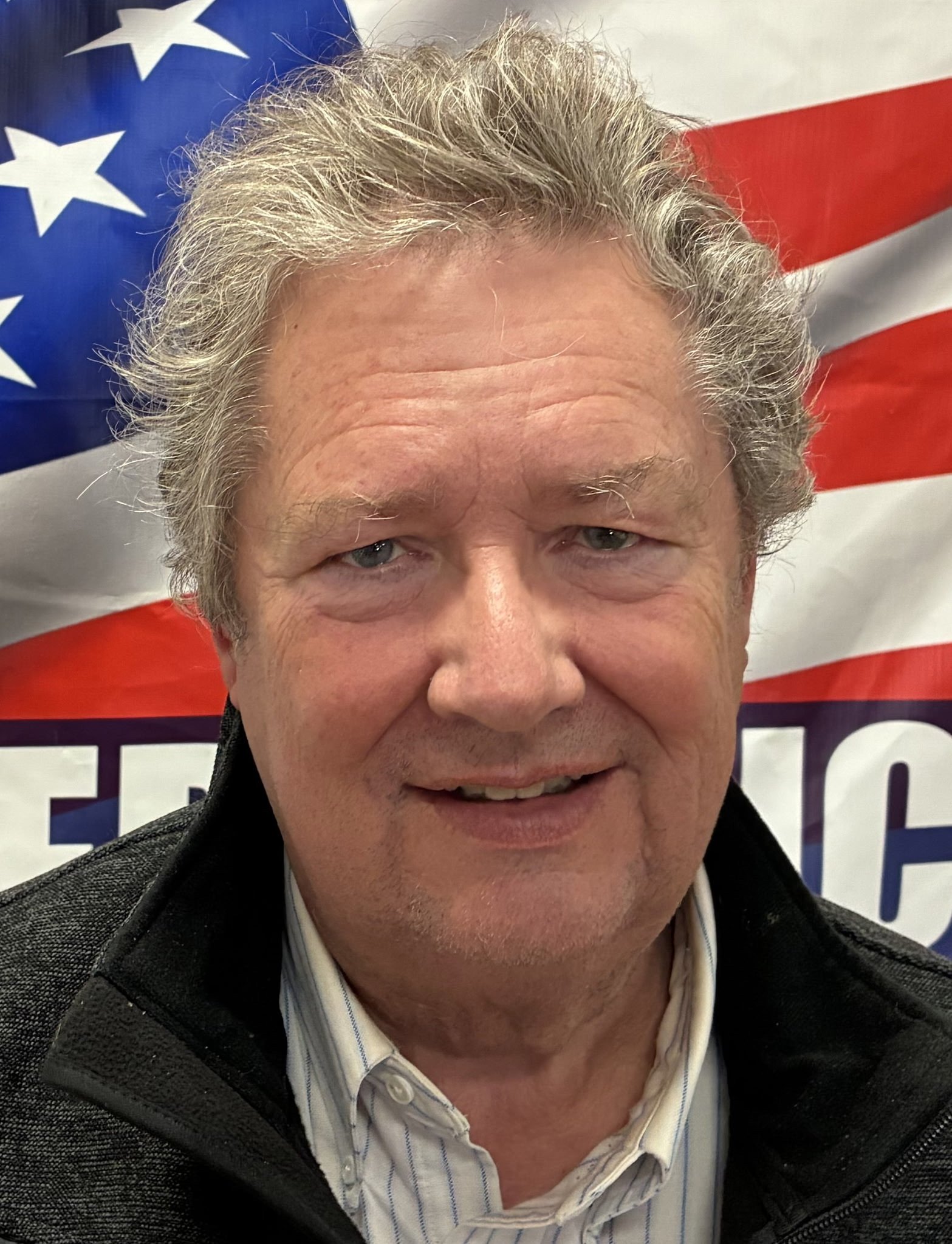 A middle-aged man with gray hair and striking blue eyes poses in front of an American flag with stars and red and white stripes.