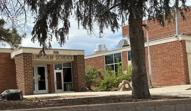 The entrance to Lincoln School, a brick building with a tree in front and shrubbery around the entrance, under a partly cloudy sky.