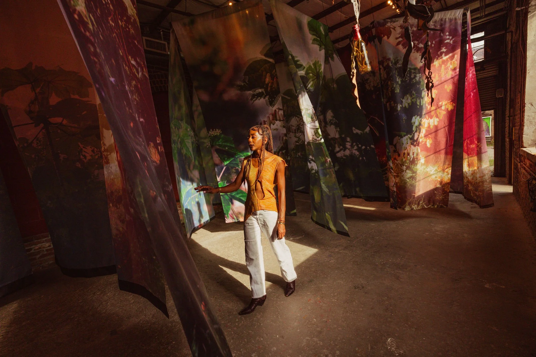 A woman with braided hair wearing a yellow top and white jeans standing in an art installation with hanging fabric panels with nature images and shadows of plants projected onto them.