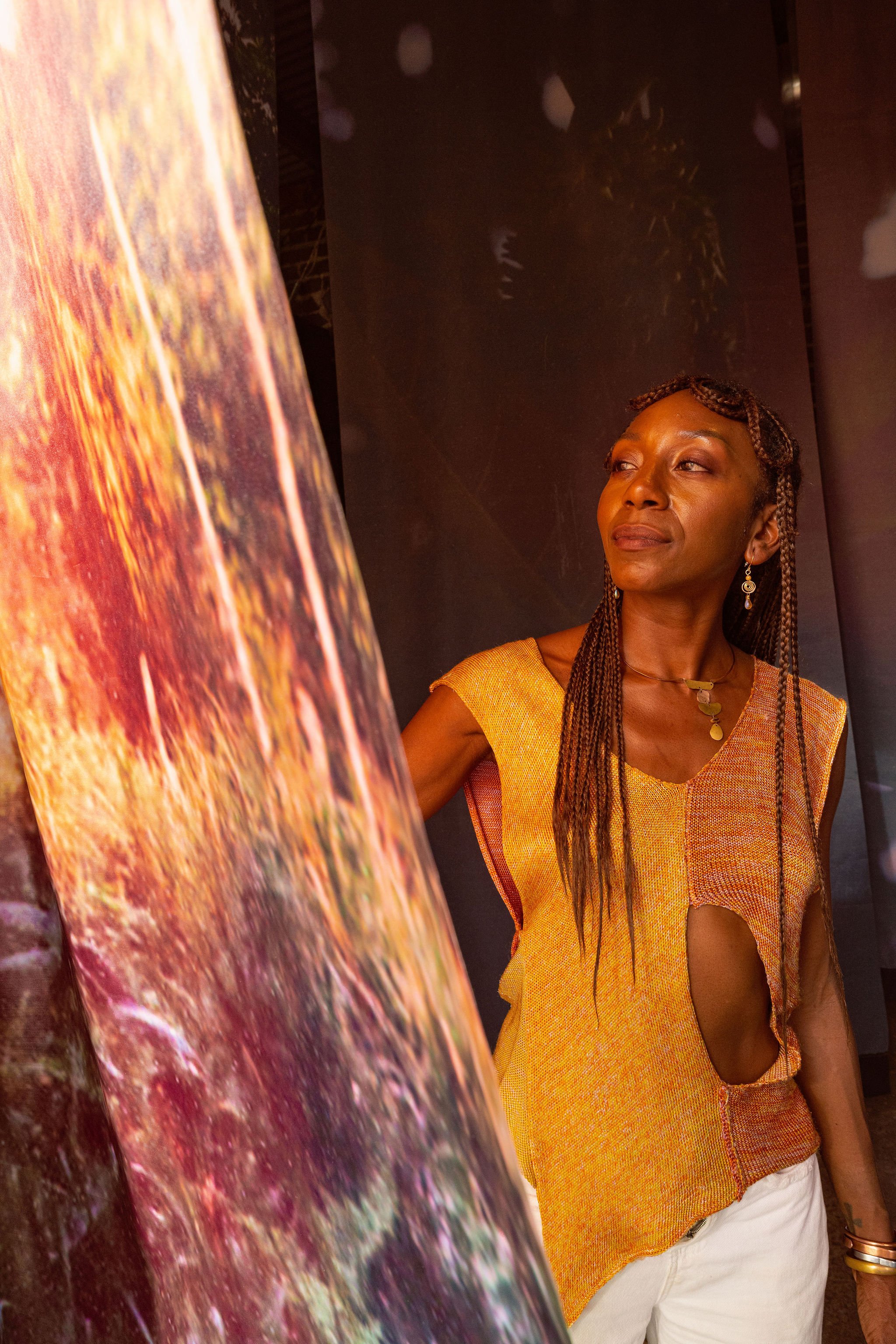A woman with braided hair and earrings, wearing a yellow top and white pants, looking at artwork in a gallery.