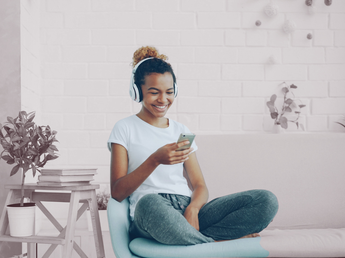 A young woman with curly hair wearing headphones, sitting cross-legged on a couch, looking at her phone and smiling.