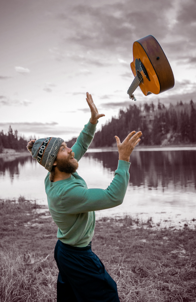 A man in a beanie and green sweatshirt is tossing a wooden acoustic guitar into the air by a lakeside with forested hills in the background.