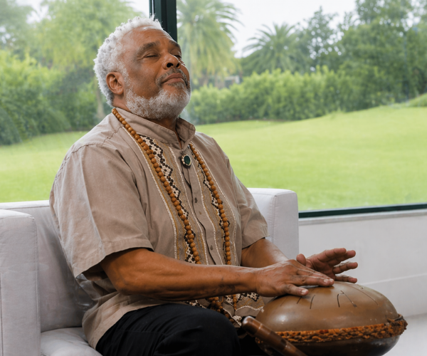 DR B Sirius, wearing traditional clothing and necklaces, meditating with hands on a singing bowl indoors with a large window showing a green outdoor landscape.