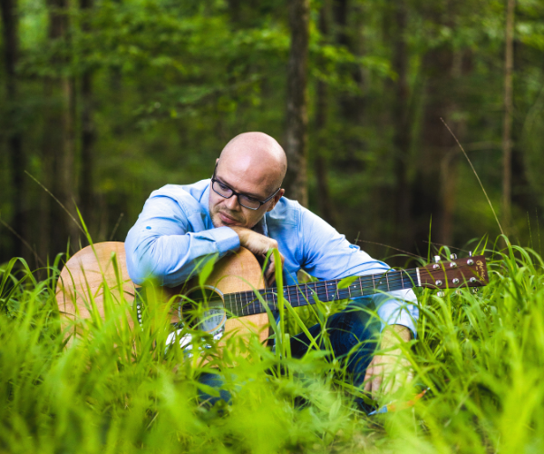 Ian Morris sitting in tall green grass playing an acoustic guitar in a forest.