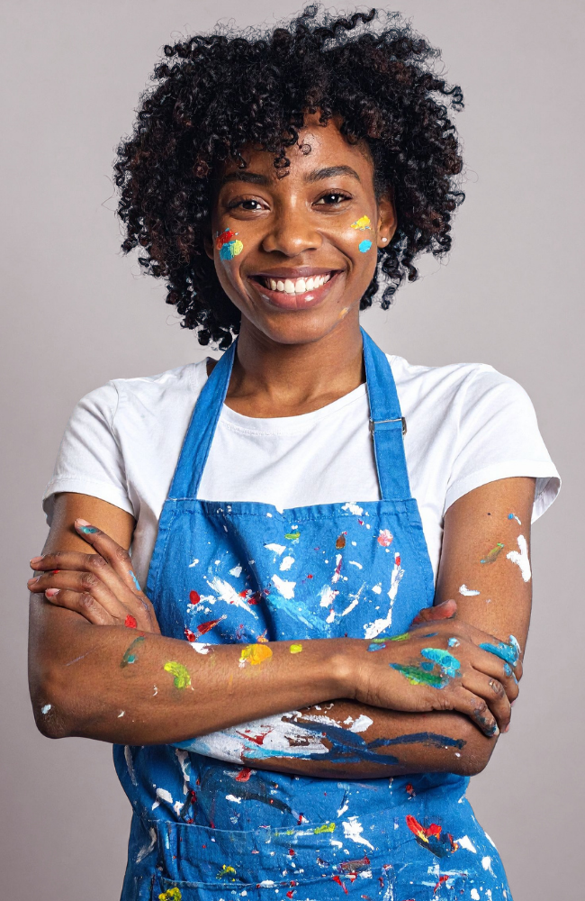 A young woman with curly black hair, smiling with arms crossed. She is wearing a white t-shirt and a blue paint-splattered apron. Her face and arms are covered in colorful paint splatters, and the background is plain gray.