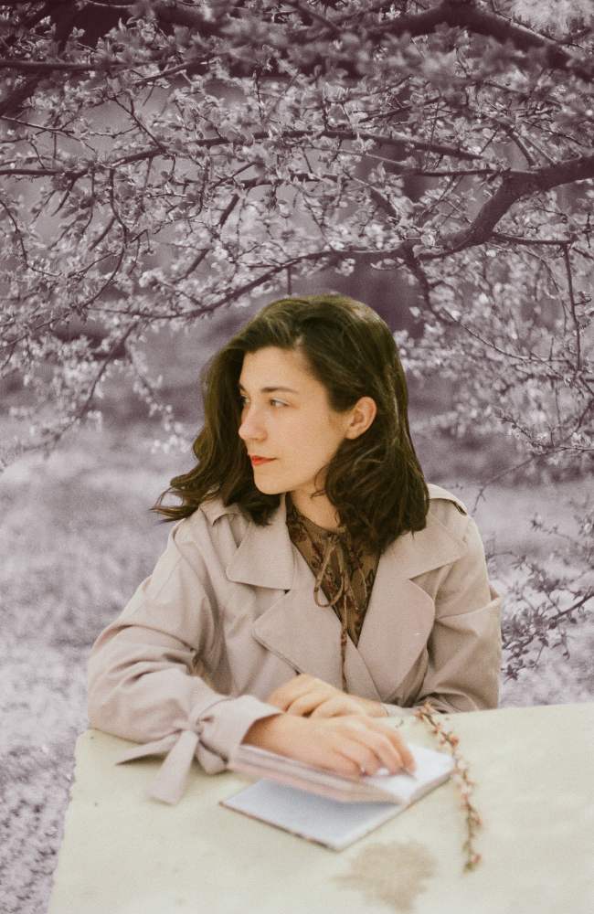 A woman with dark wavy hair, wearing a beige trench coat and patterned blouse, sitting at a table with an open notebook in front of her, outdoors with blooming tree branches in the background.