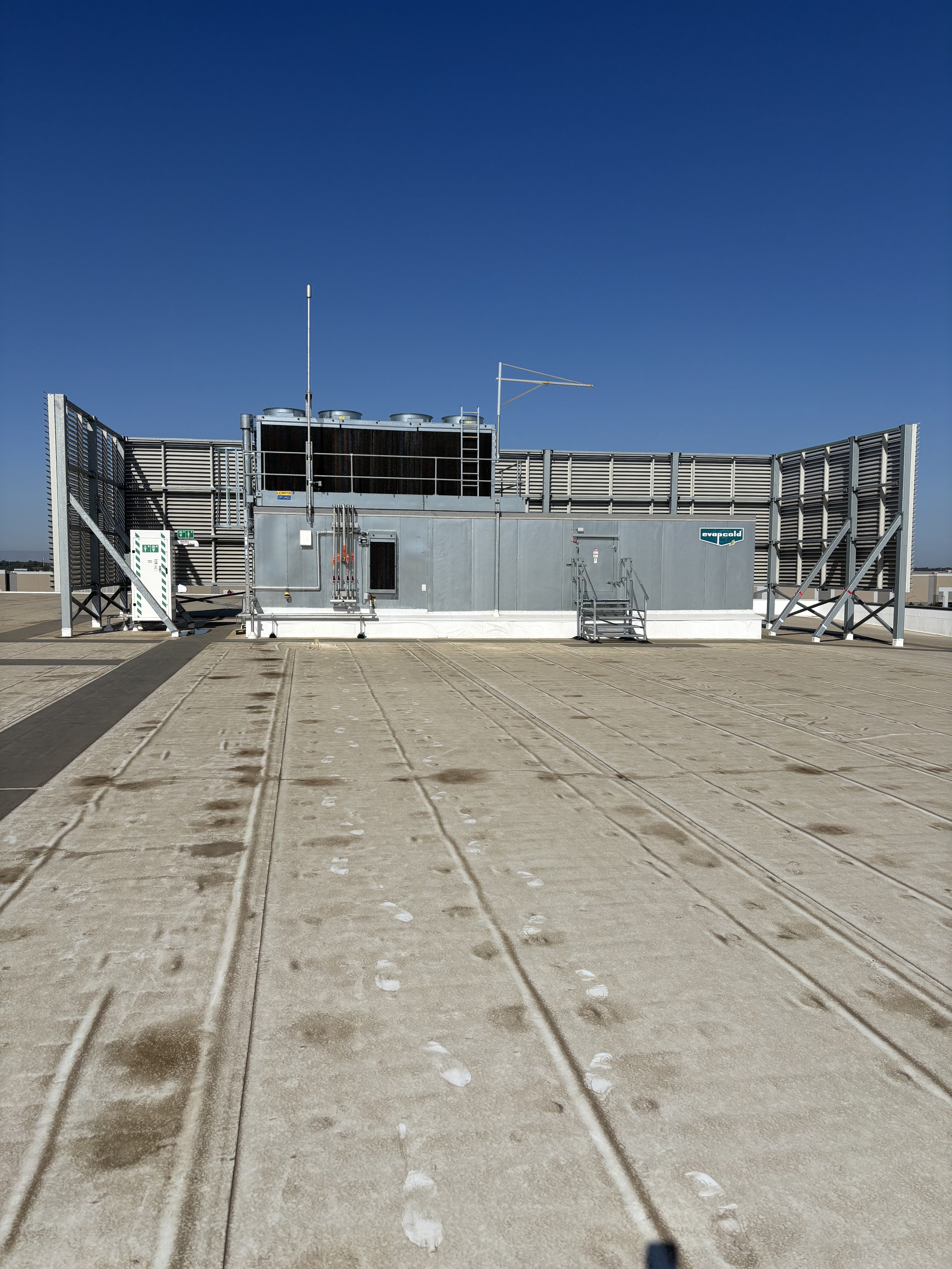Rooftop HVAC unit with metal exterior, fans, antennas, and safety railings under a clear blue sky.