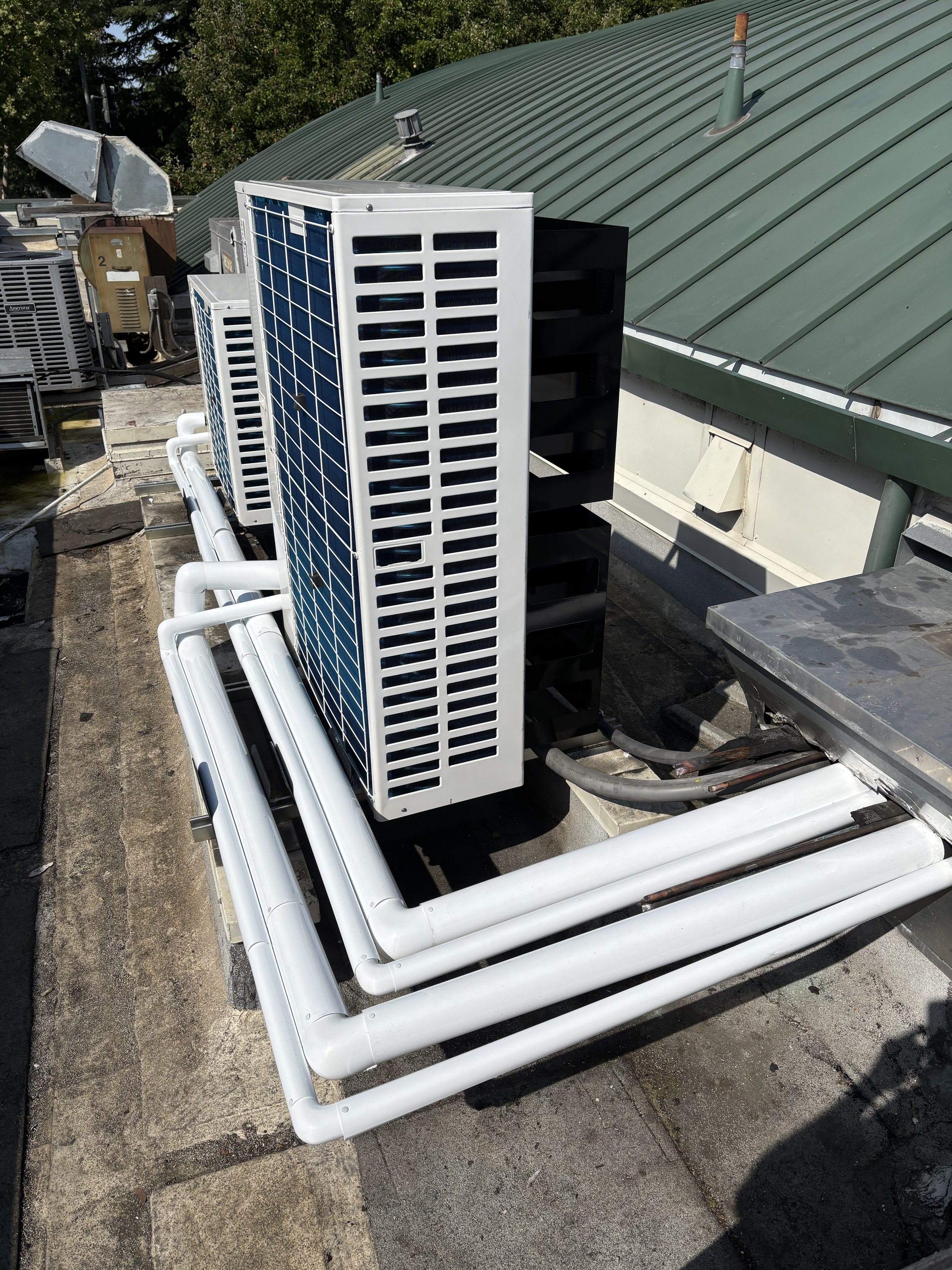 Multiple air conditioning units and white PVC pipes on a rooftop with a green metal roof.