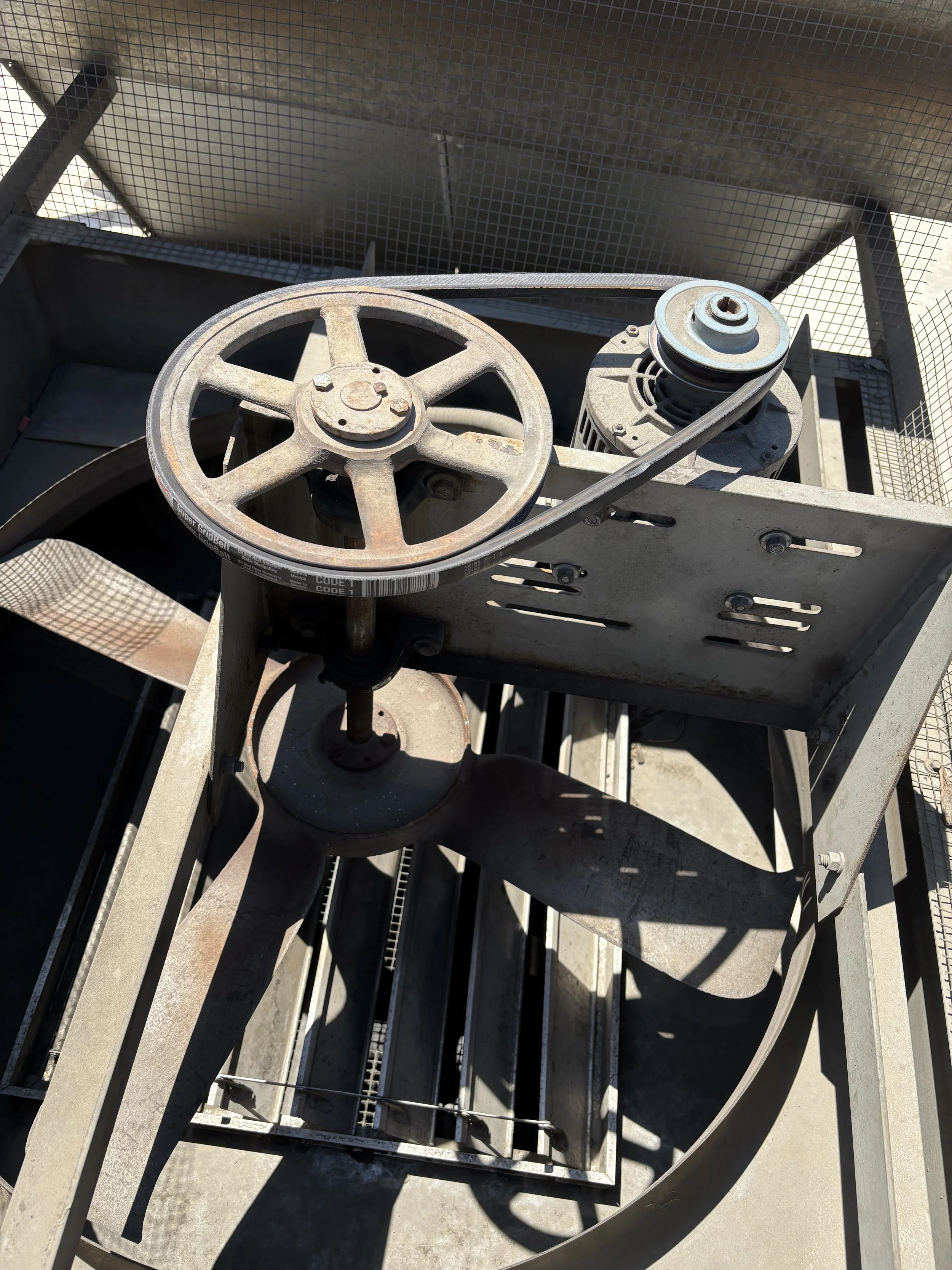 A metal outdoor pizza oven with a metal wheel and belt on the front. The oven has a wire mesh surround and is situated on a metal stand with slats and rods.