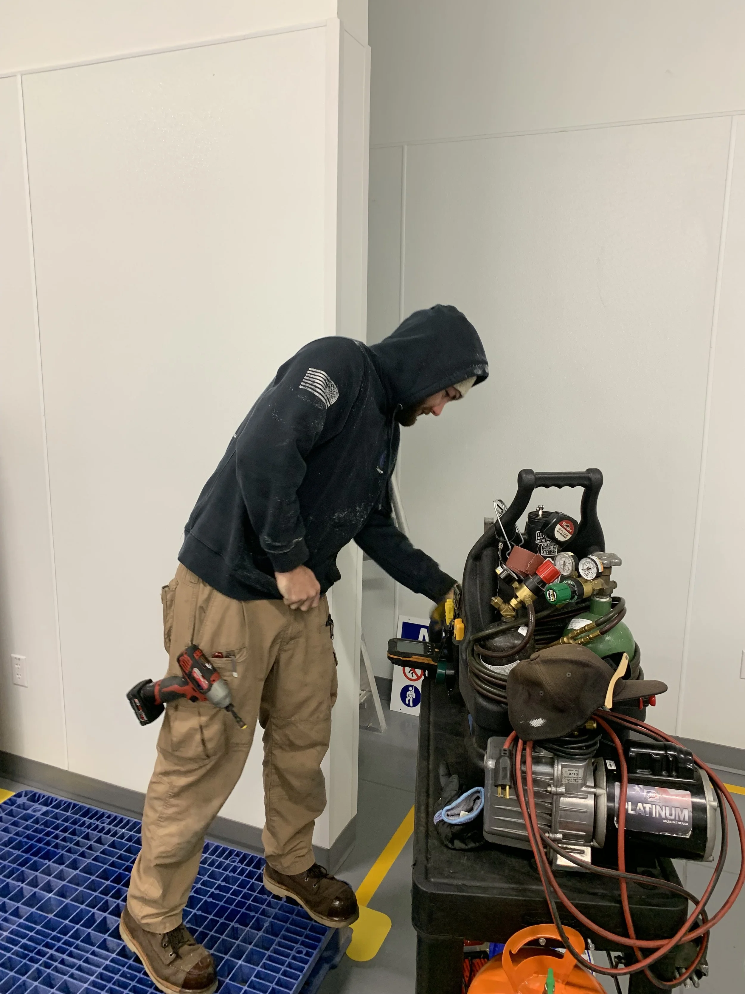 A man wearing a black hoodie and beige work pants working with HVAC or refrigeration tools in an indoor utility room or maintenance area, next to a cart filled with gauges, hoses, and other equipment.