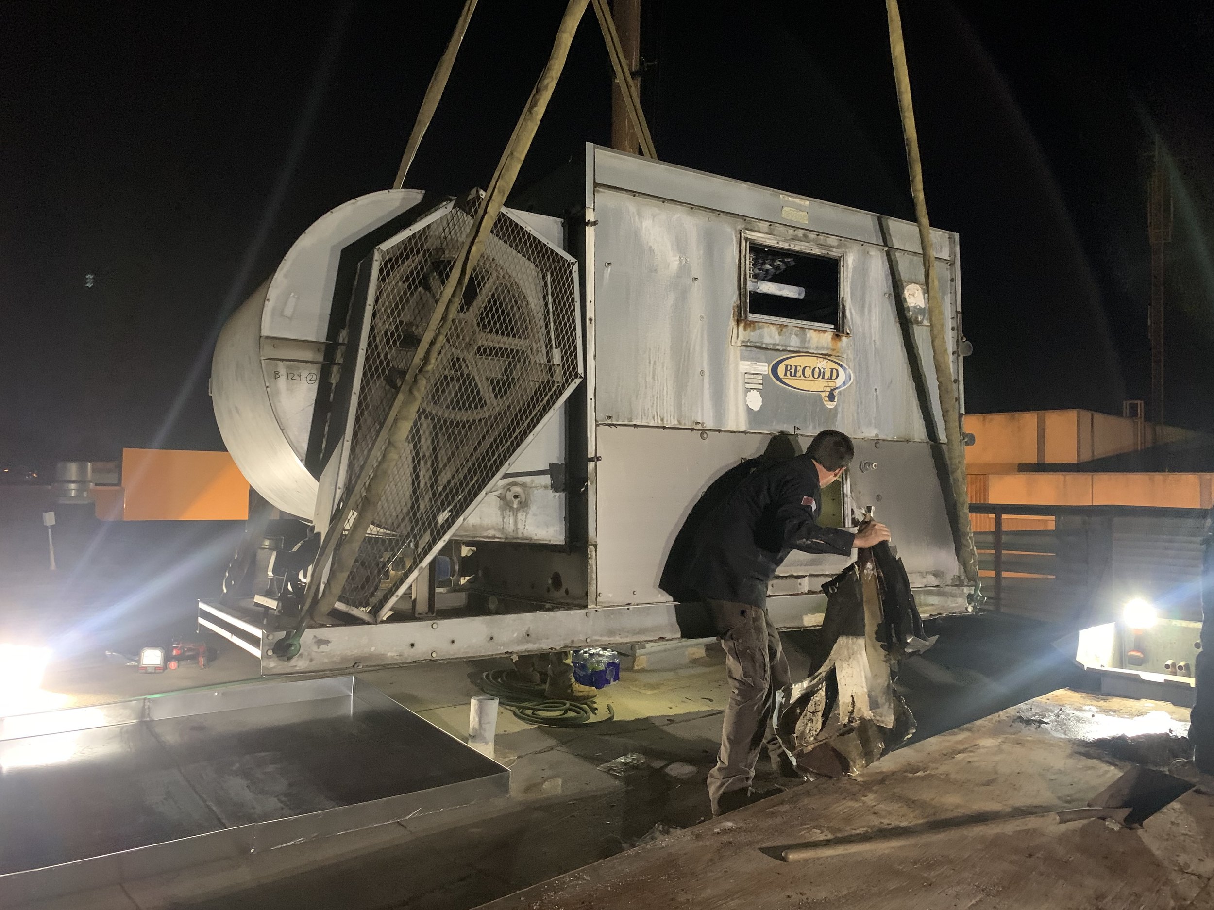 Construction workers are installing a large metallic HVAC or refrigeration unit outdoors at night. The unit has a fan on one side and a small window or opening, with tools and equipment around them.