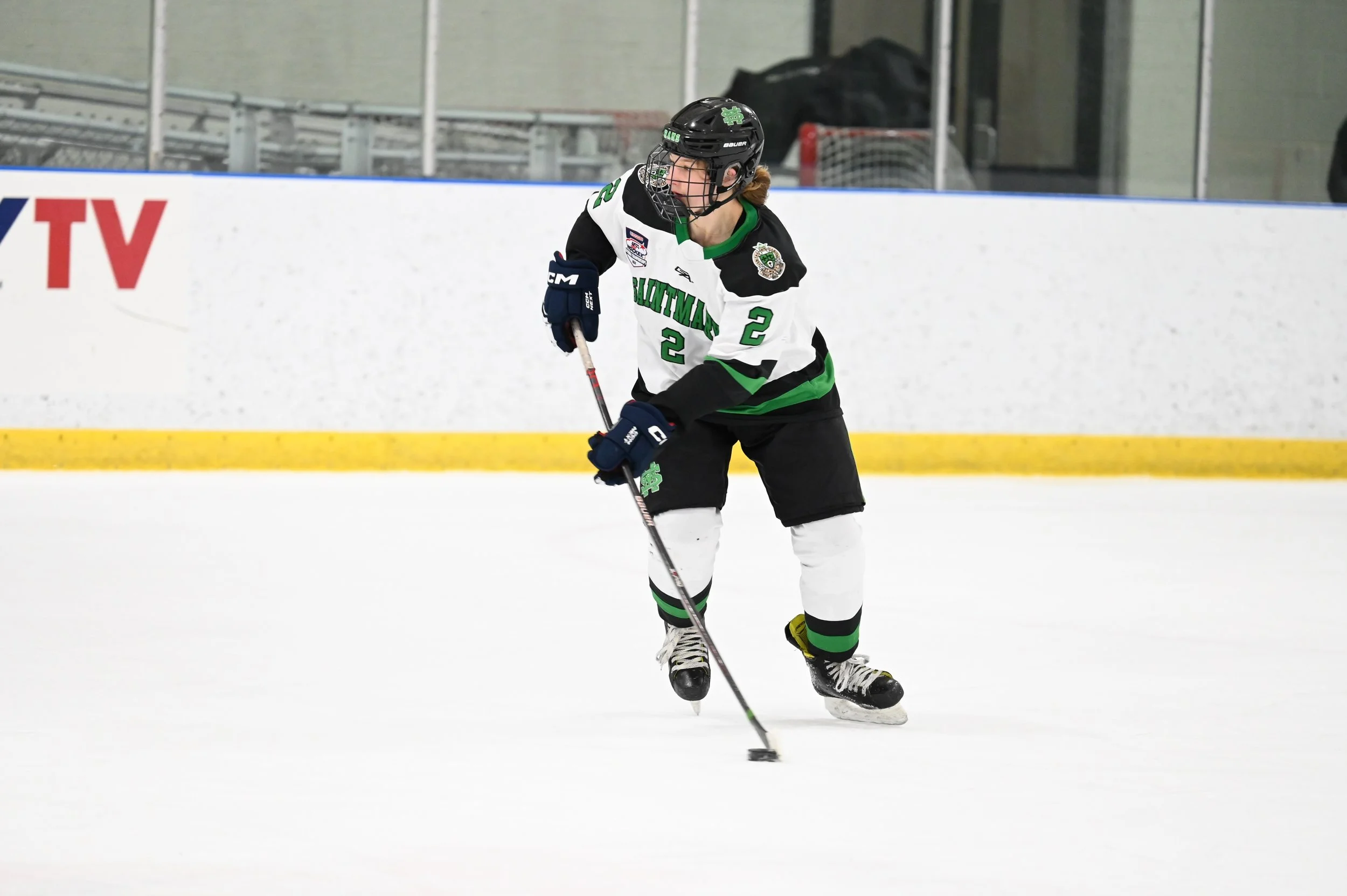 A female hockey player in a black, white, and green jersey with the number 22, wearing a black helmet and gloves, is skating on ice and handling a hockey puck.
