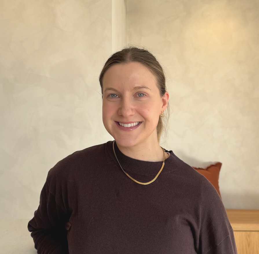 Smiling woman with blue eyes and light brown hair in a casual black top, wearing a gold necklace and earrings, sitting indoors against a light-colored wall with a brown pillow in the background.