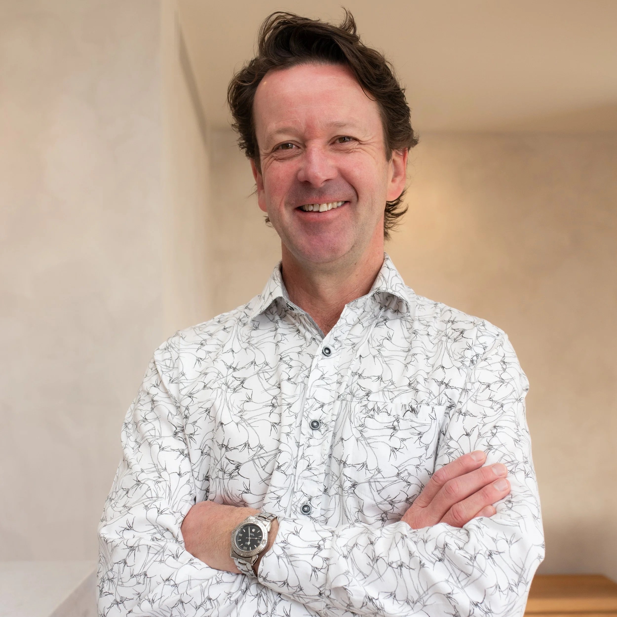 A smiling man with dark, wavy hair wearing a light-colored shirt with a subtle leaf pattern, standing against a plain, light-colored wall.