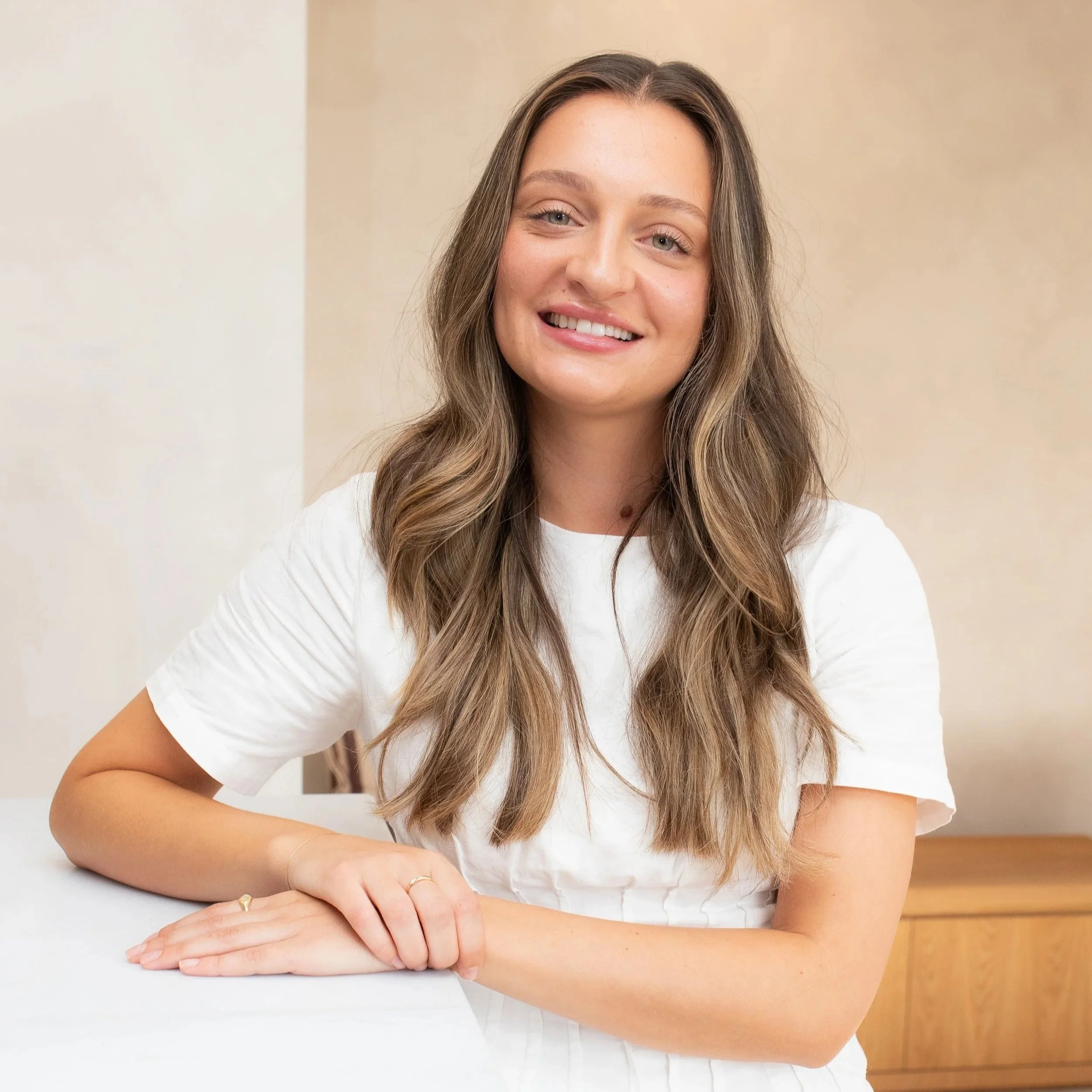 A young woman with long brown wavy hair, light skin, wearing a striped button-up shirt, smiling with visible teeth, standing against a plain light-colored background.