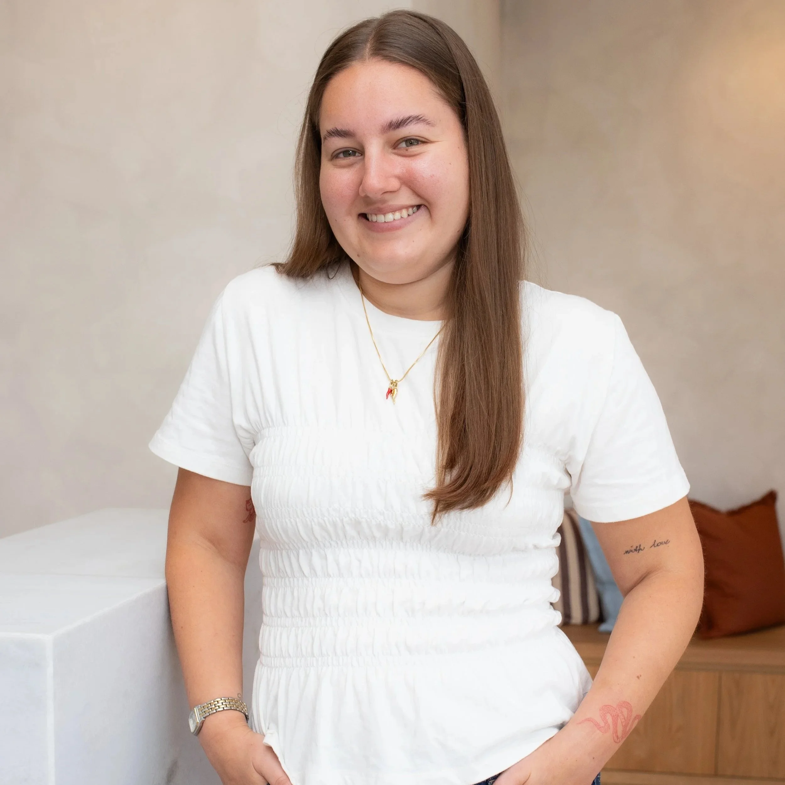 A young woman with long brown hair, smiling and wearing a sleeveless yellow top with buttons, standing against a plain white background.