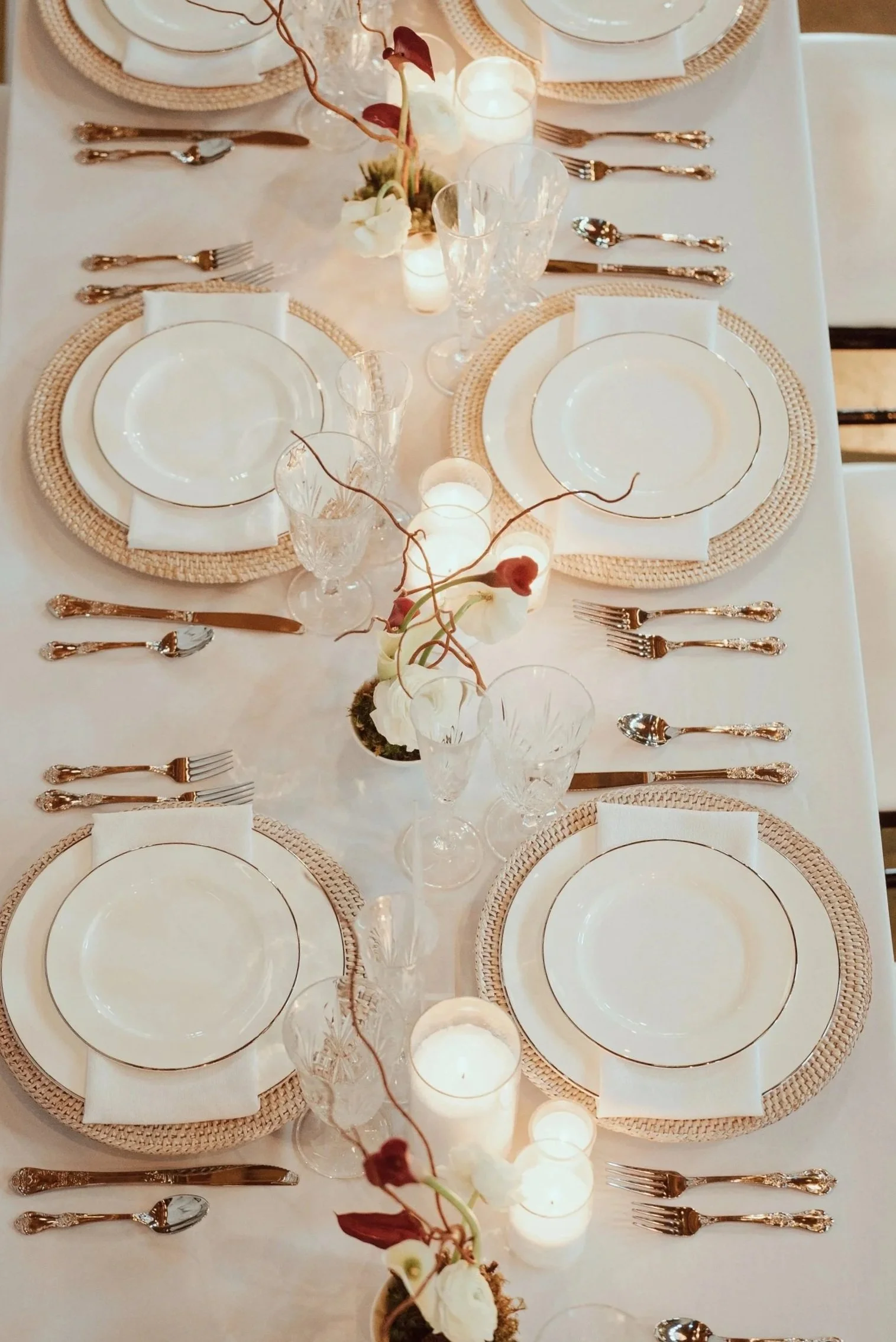 Elegant dining table setup with white plates, silverware, clear glassware, and wine glasses. Centerpiece includes flowers and candles, with woven placemats on a white tablecloth.