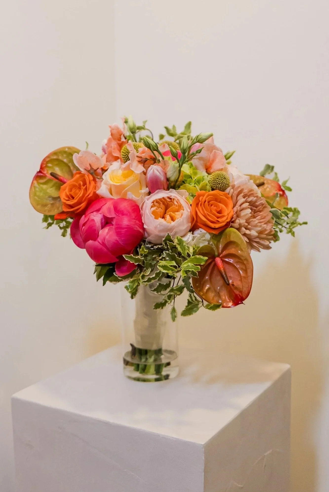 Arrangement of various pink, orange, yellow, and green flowers in a glass vase on a white pedestal against a light background.