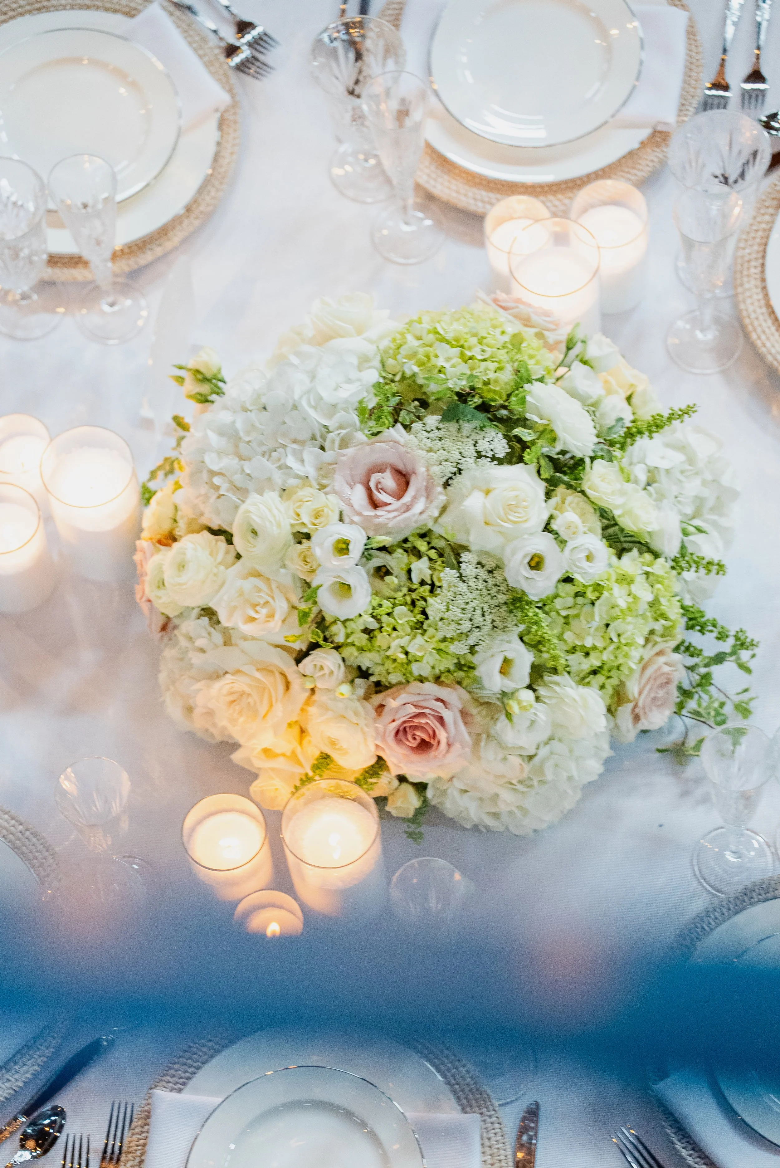 A formal dining table decorated with a large pastel-colored floral centerpiece, surrounded by white plates, silverware, clear glassware, and lit candles, set for a special occasion or dinner event.