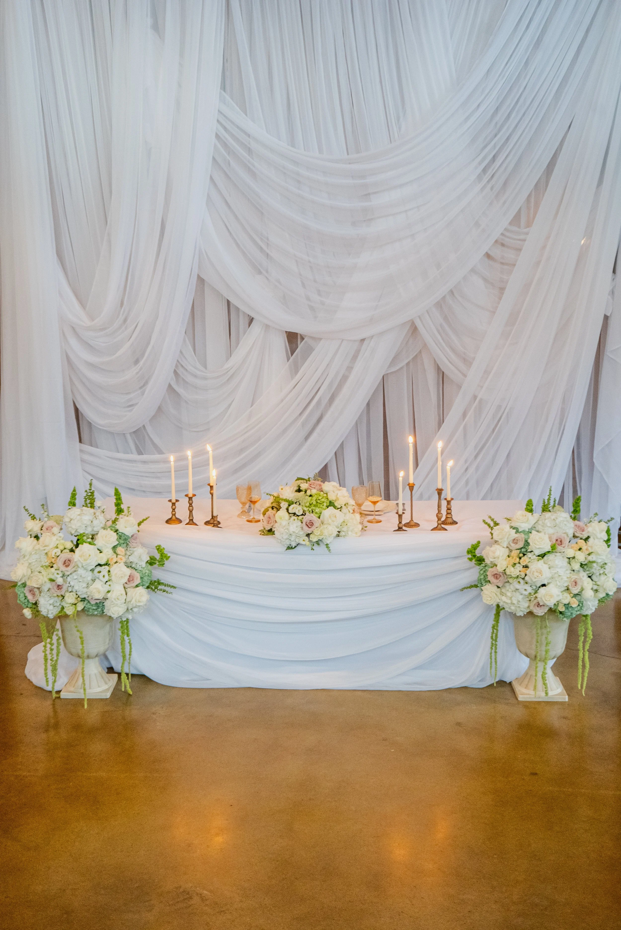 Elegant wedding reception table with white draped fabric backdrop, floral centerpieces, candle holders, and table settings.