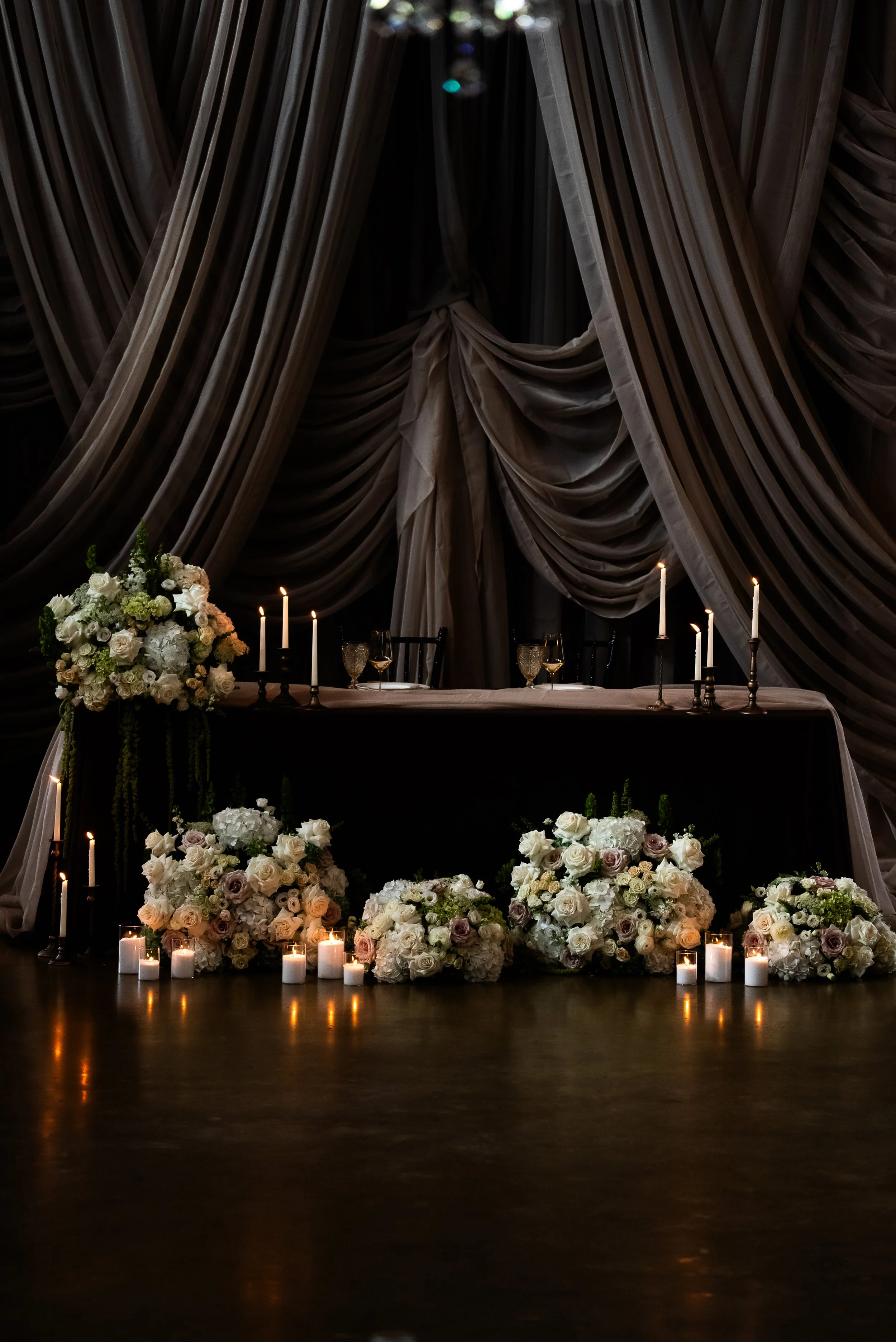 A decorated table with multiple floral arrangements of white, cream, and blush roses, surrounded by lit candles in glass holders, set against a backdrop of large draped curtains and dim lighting.