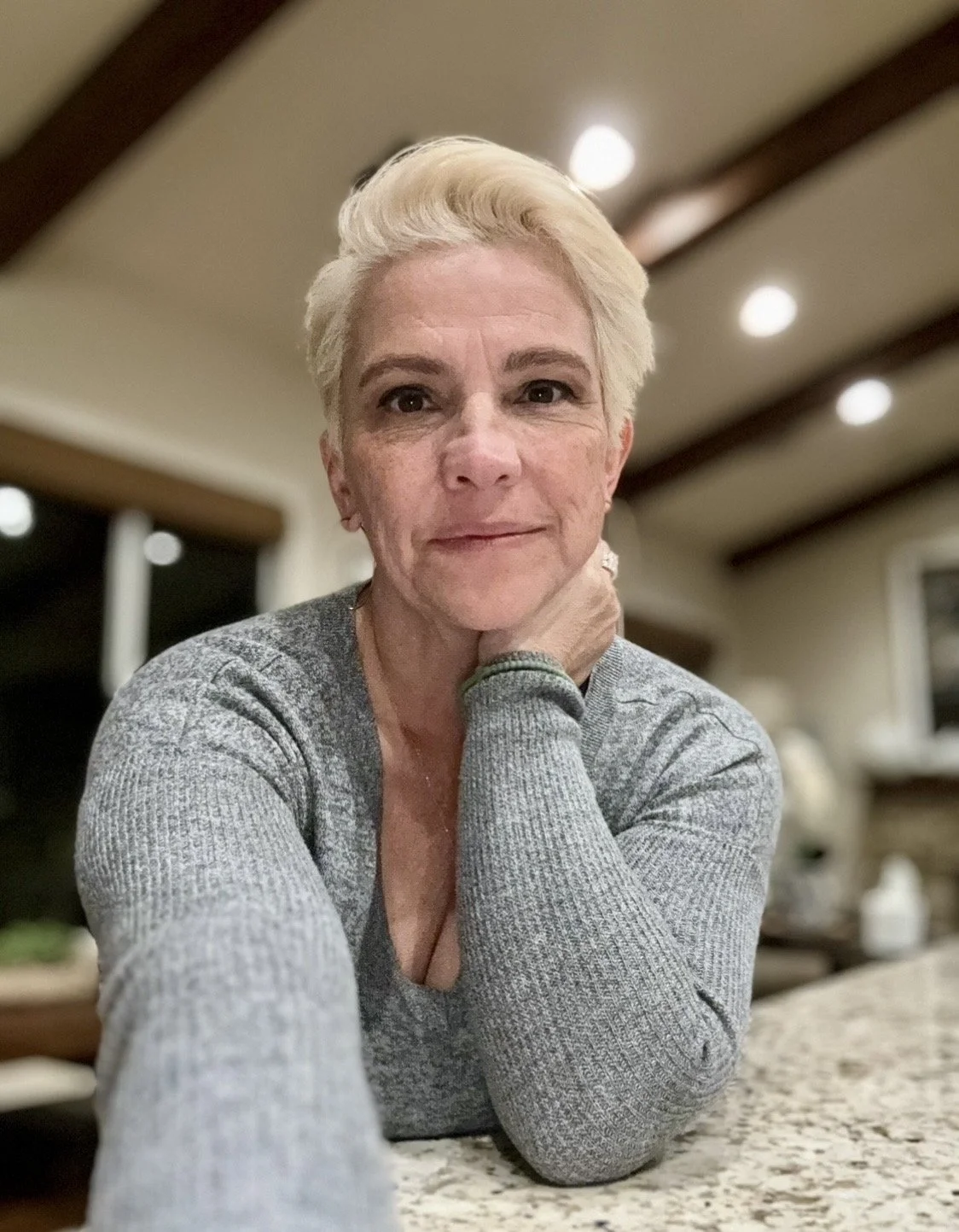 A middle-aged woman with short blonde hair sitting at a kitchen counter, resting her chin on her hand, looking into the camera.