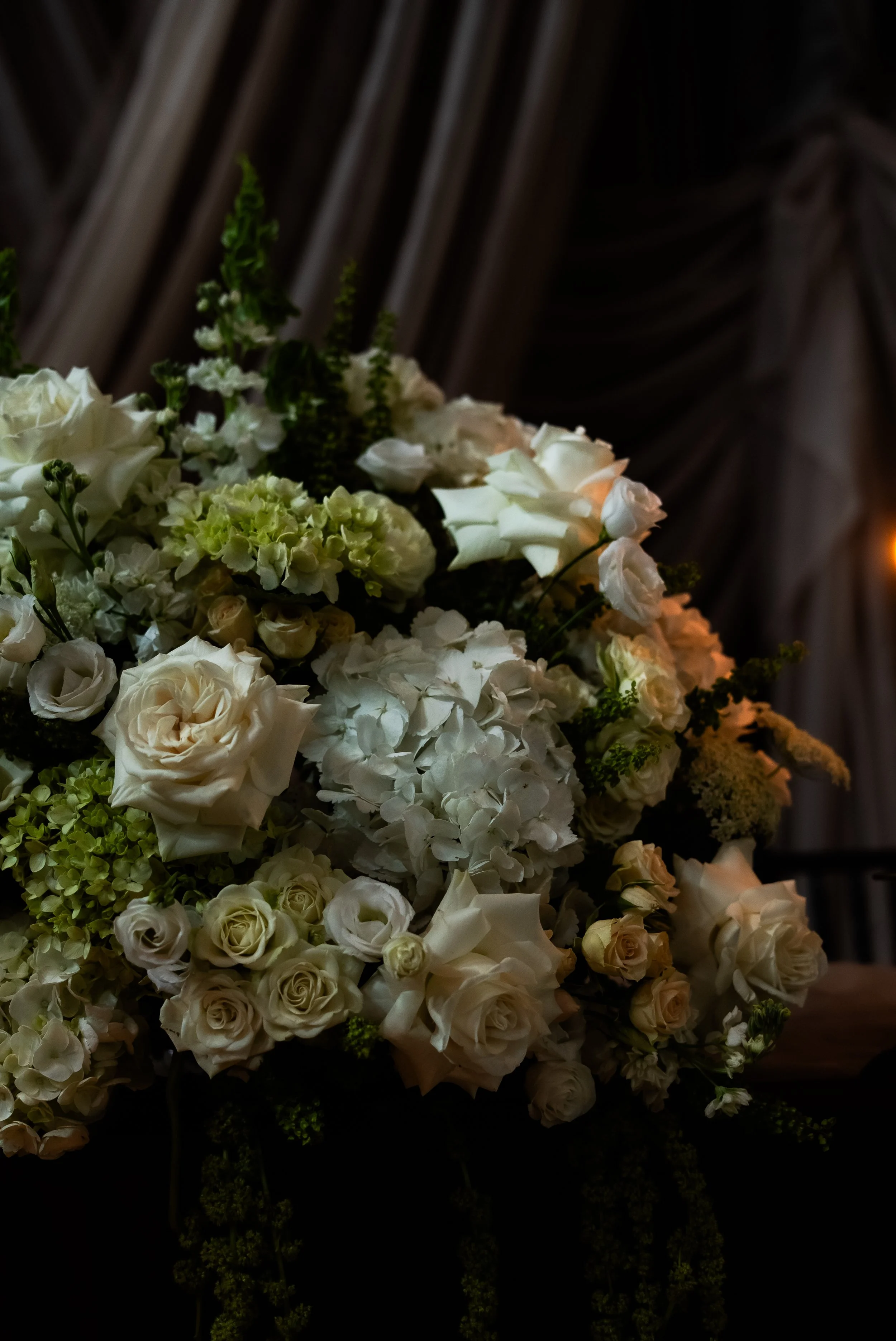 A large bouquet of white and cream-colored flowers, including roses and hydrangeas, arranged with greenery, against a dark fabric backdrop.