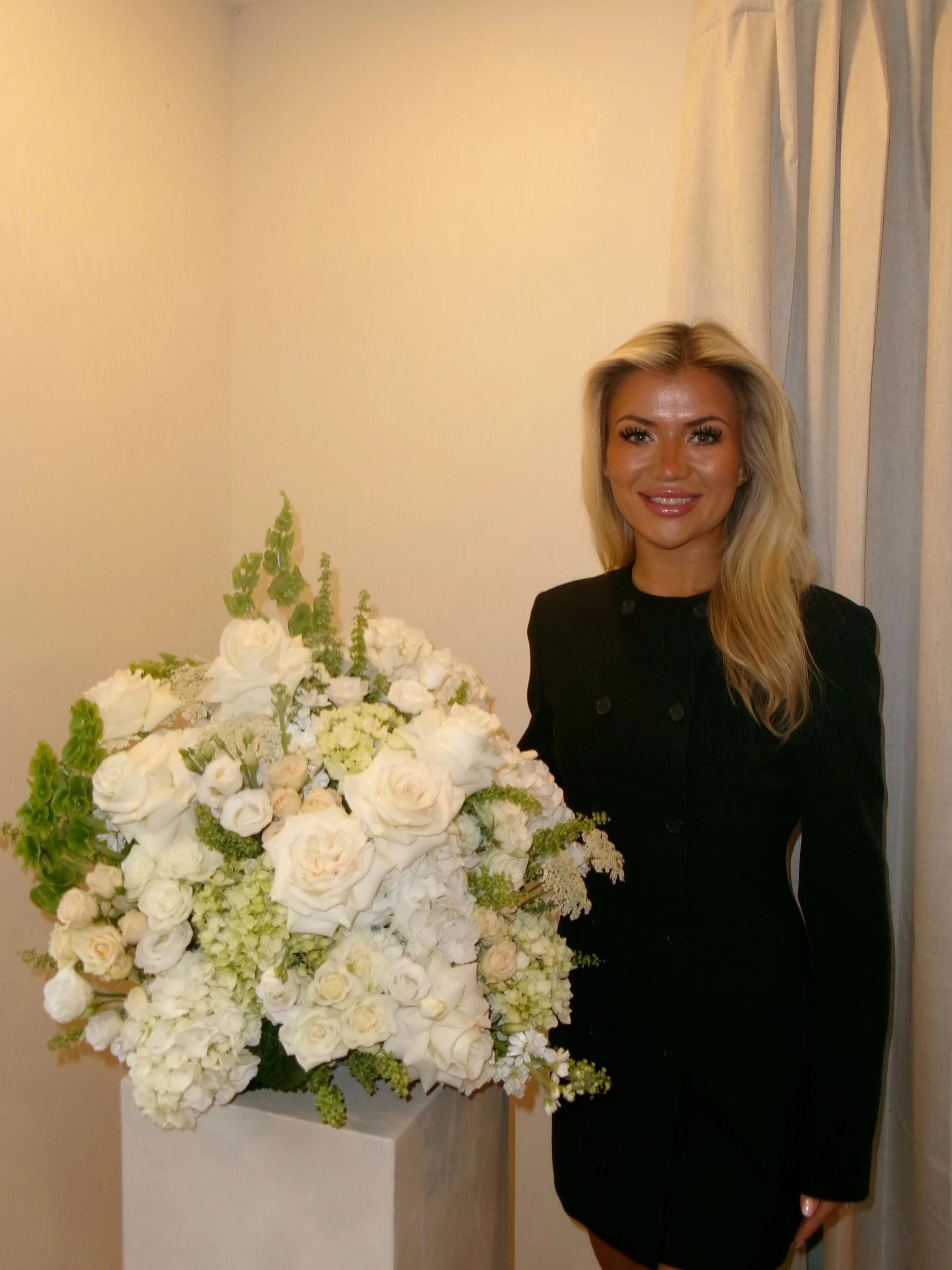 A woman with blonde hair wearing a black dress standing beside a large bouquet of white flowers on a white pedestal, beige wall, and curtain in the background.