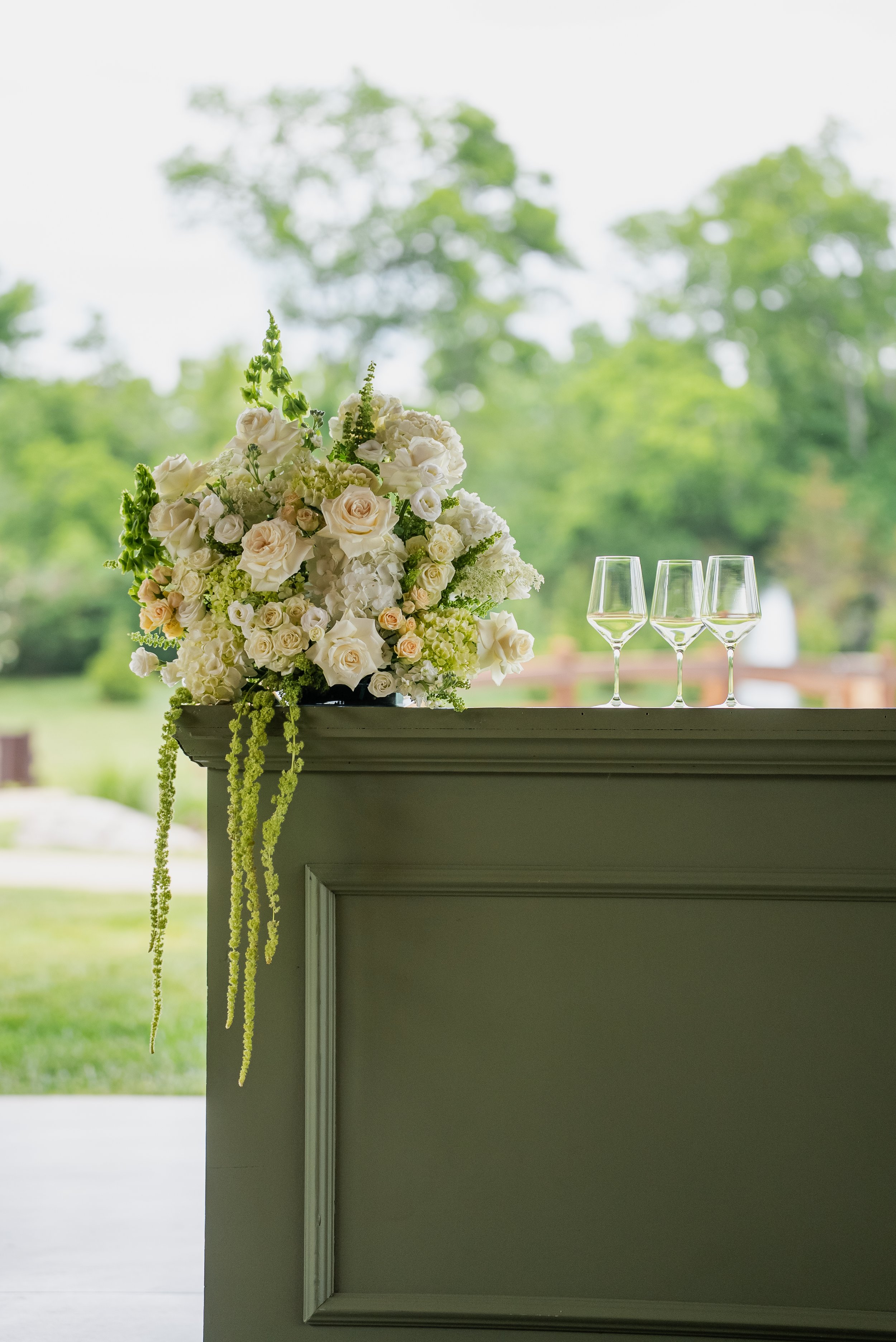 A floral arrangement with white and cream roses, green buds, and hanging green amaranth, placed on a gray piece of furniture. Three wine glasses filled with white wine are on the right side of the arrangement. The background shows a blurry outdoor sc