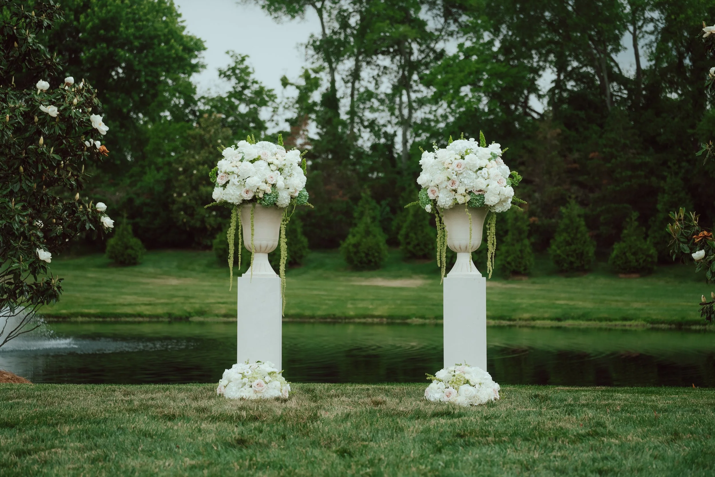 Two large white flower arrangements on tall white pedestals near a body of water, surrounded by green trees and grass.