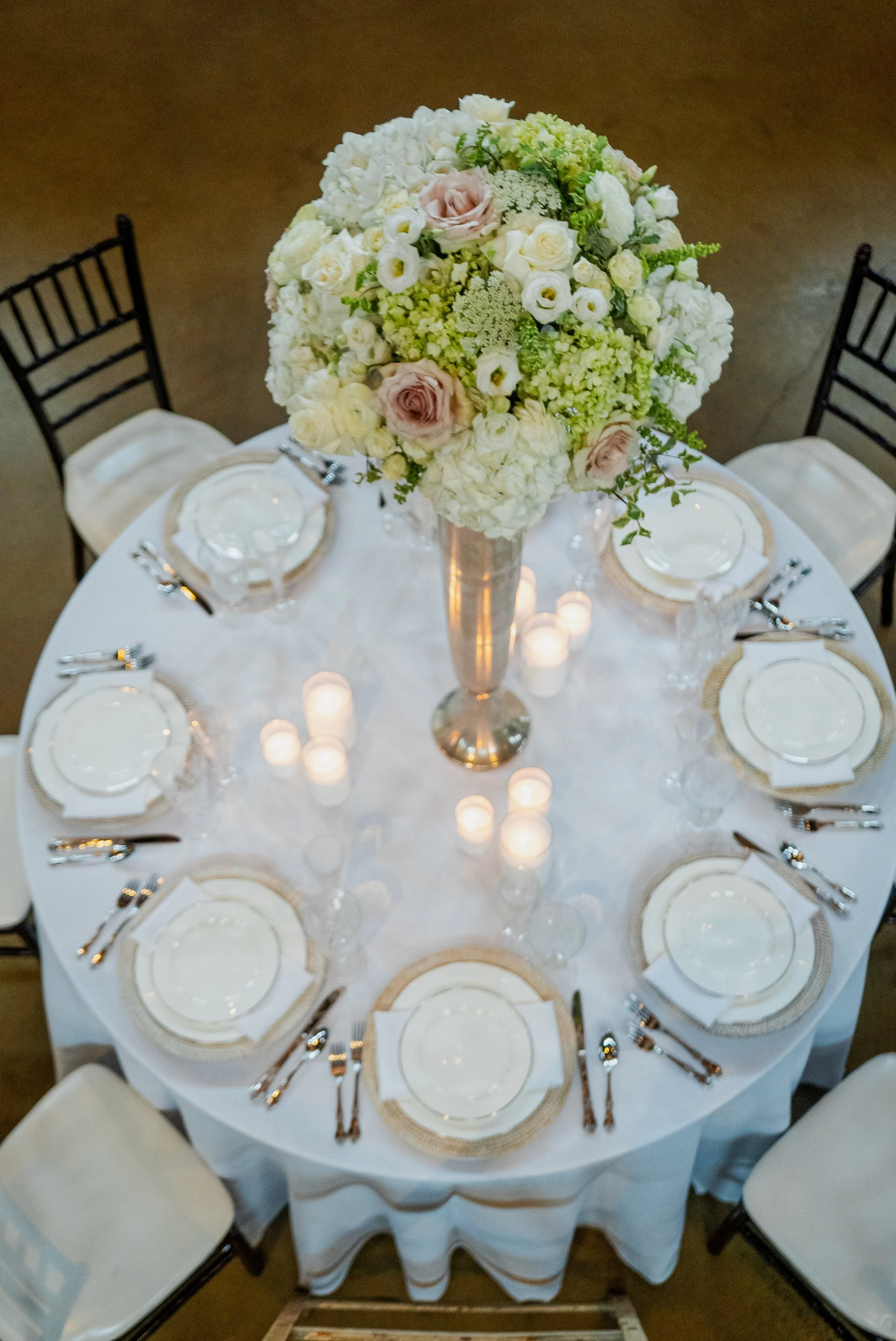 Elegant round table set for a formal event with a white tablecloth, surrounded by cream-colored chairs. The center features a large floral arrangement with white, cream, and pale pink flowers. Small candles are scattered around the centerpiece, and p