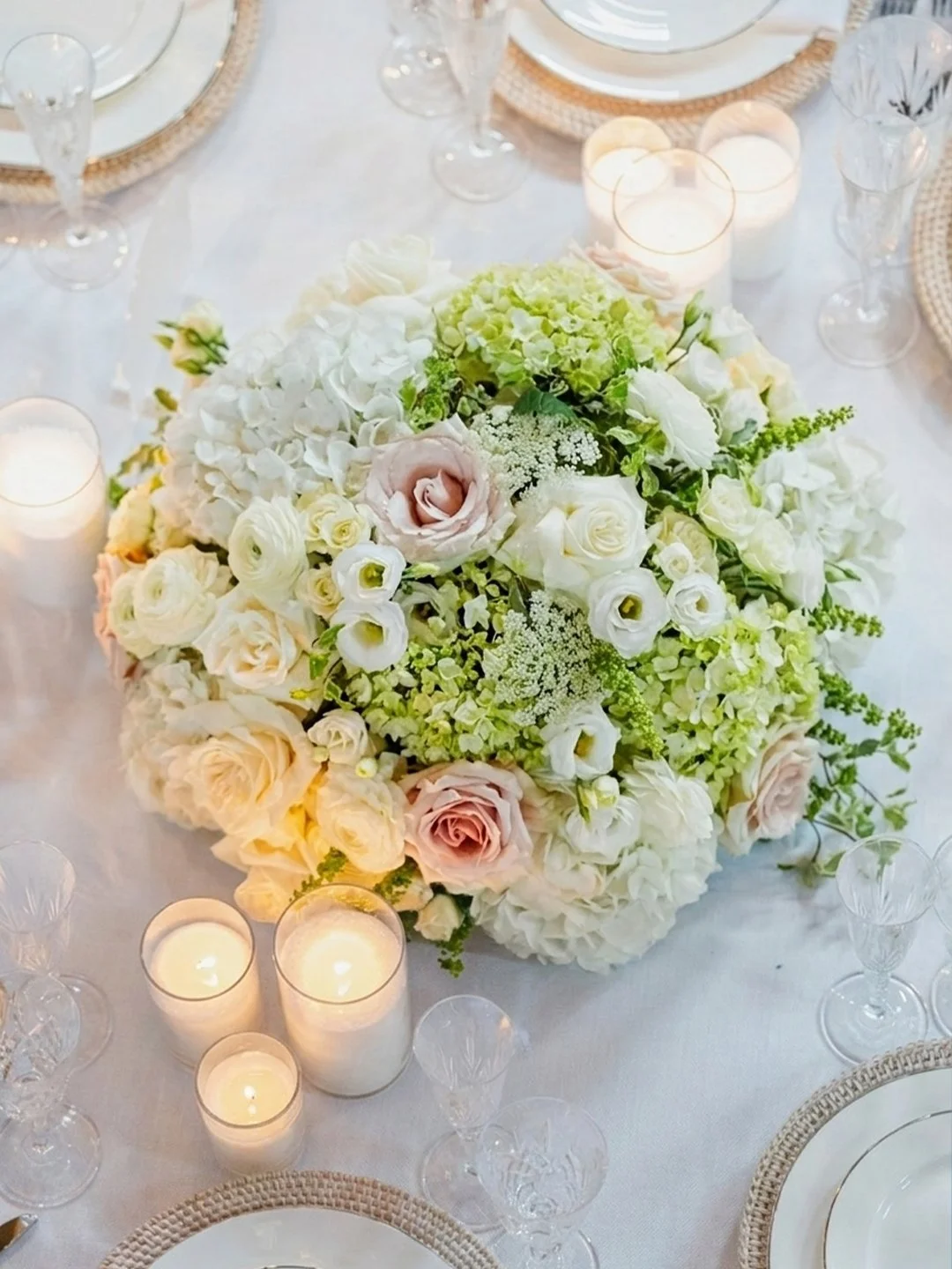 A table centerpiece with white, blush pink, and cream-colored flowers including roses, hydrangeas, and lisianthus, surrounded by lit candles and elegant tableware.