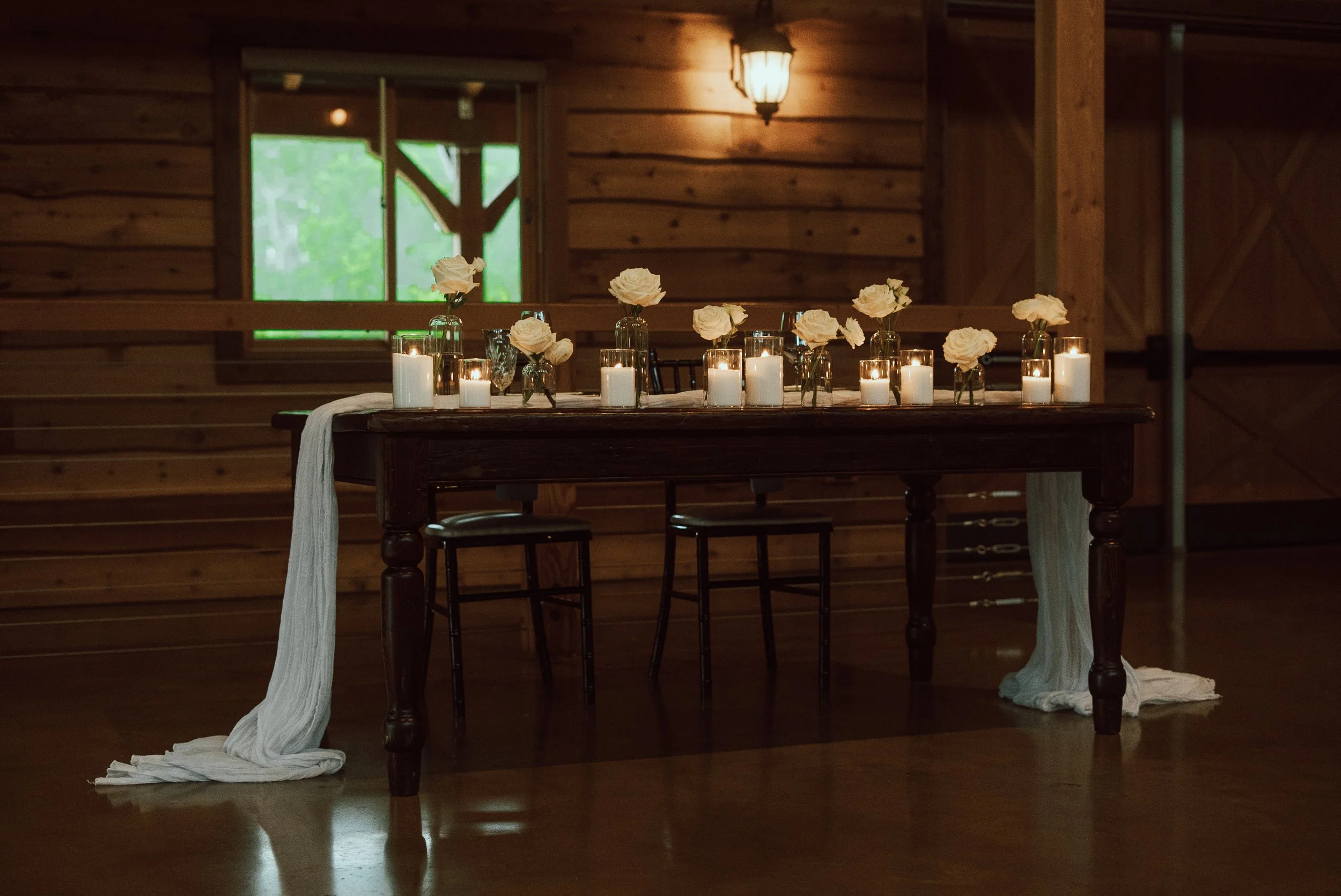 A wooden table decorated with white roses in glass vases and lit candles, set inside a rustic wooden room with a window and warm lighting.