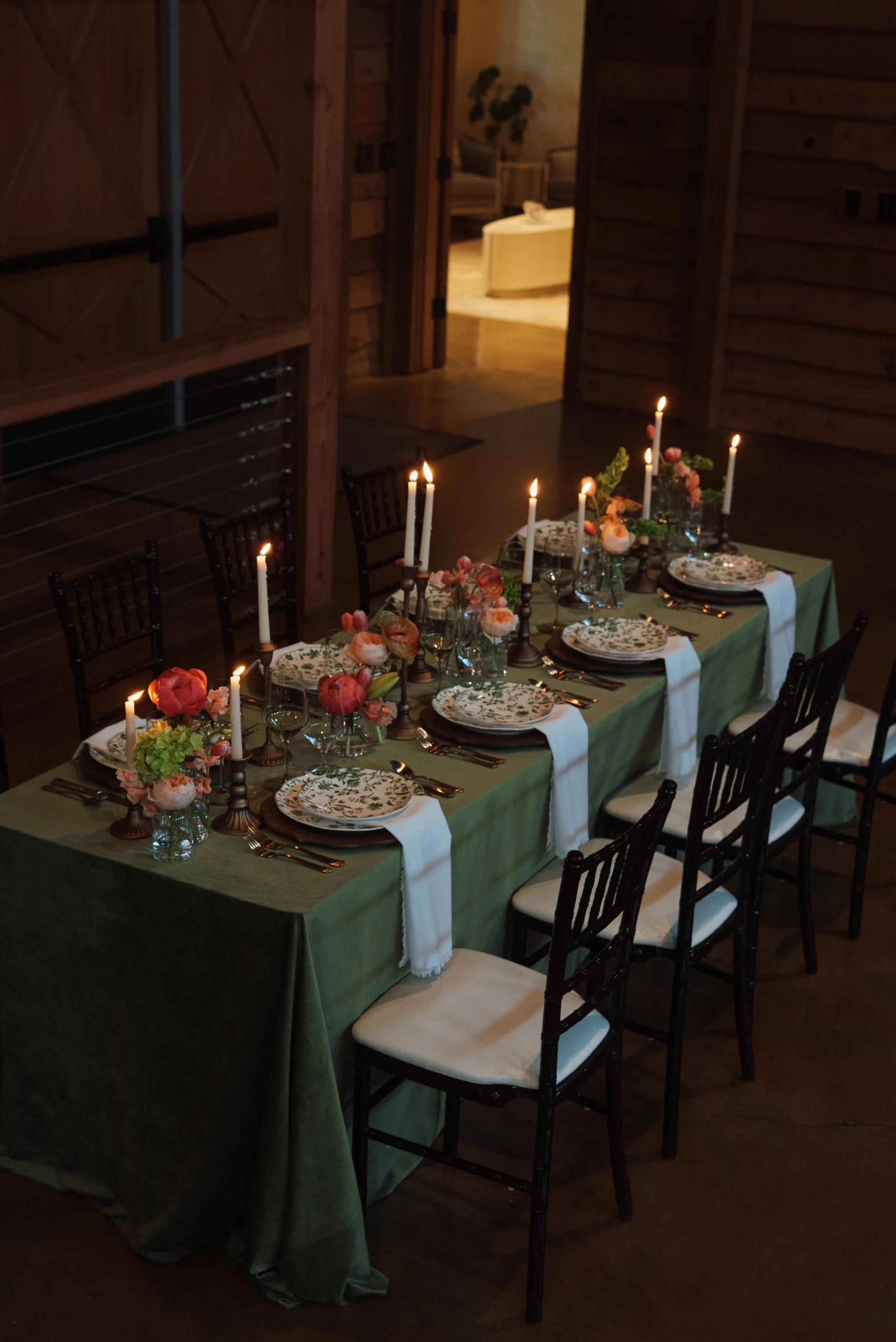 A long dining table set for a meal with floral and candle decor in a rustic room with wooden walls and a view into an adjacent room.