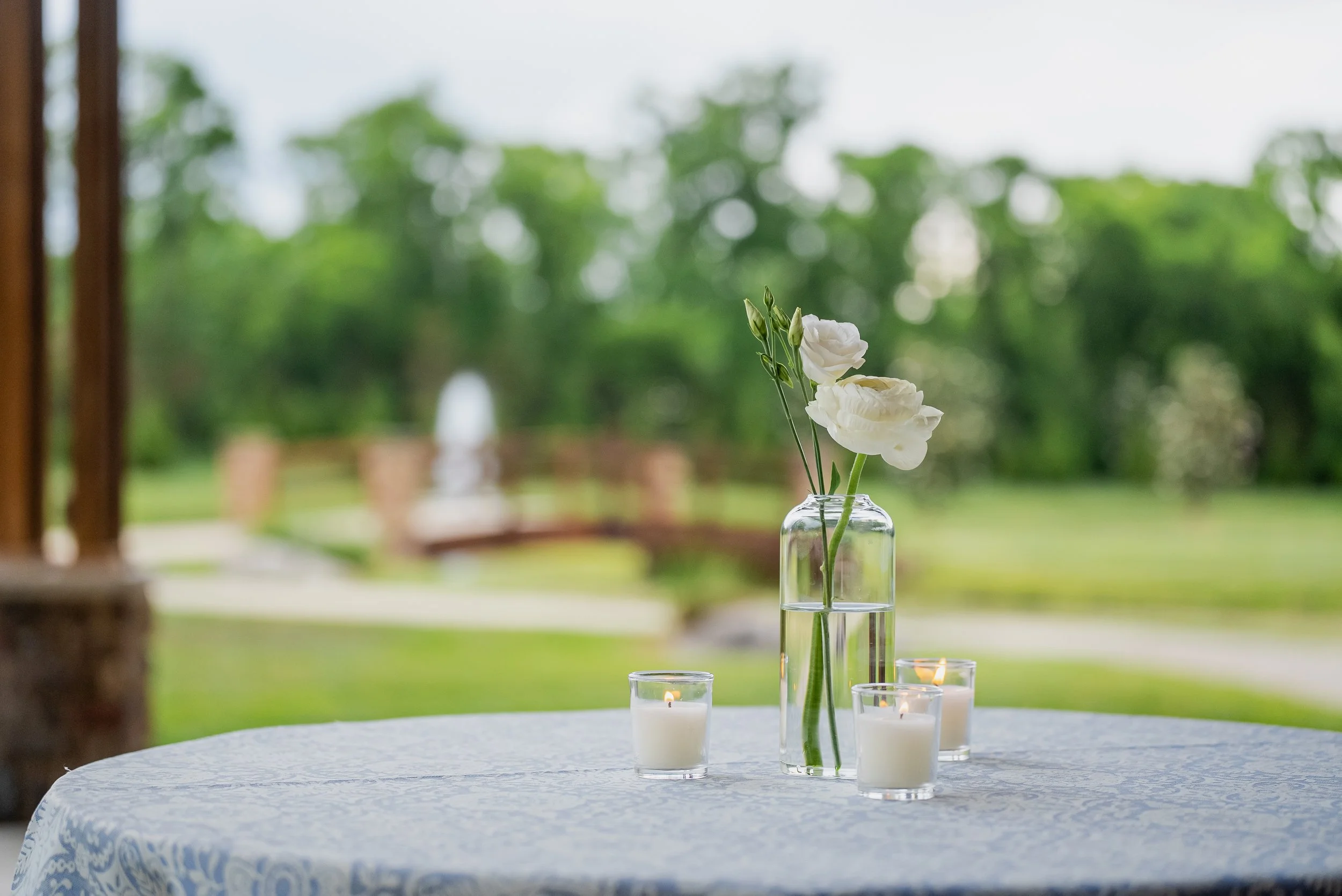 A table with a white patterned tablecloth holding a clear glass vase with white flowers and three small white candles in glass holders, set outdoors with green trees in the background.