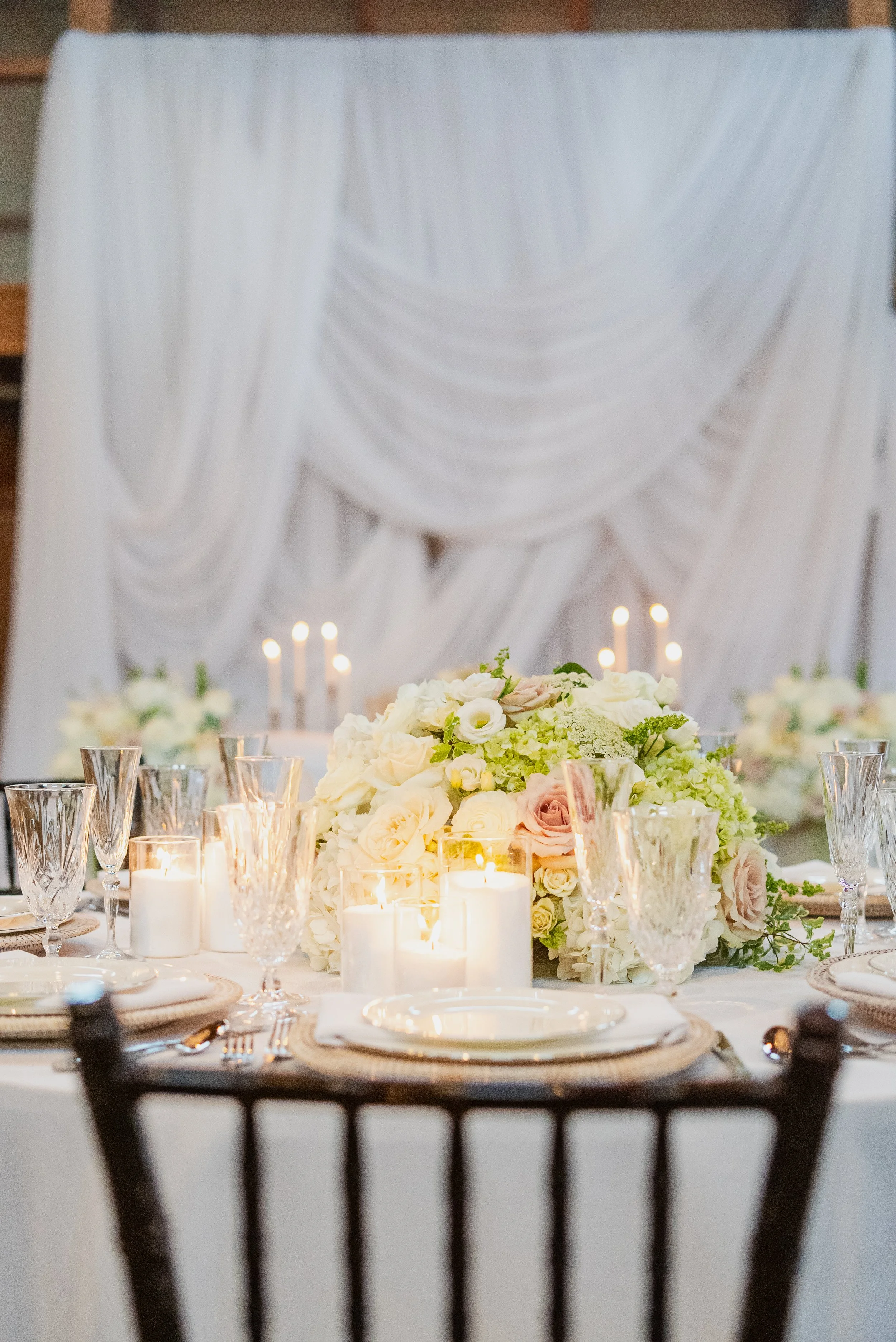 A table decorated for a wedding or formal event with a large floral centerpiece of white and blush roses, surrounded by candles and glassware, with a backdrop of white drapery and candles.