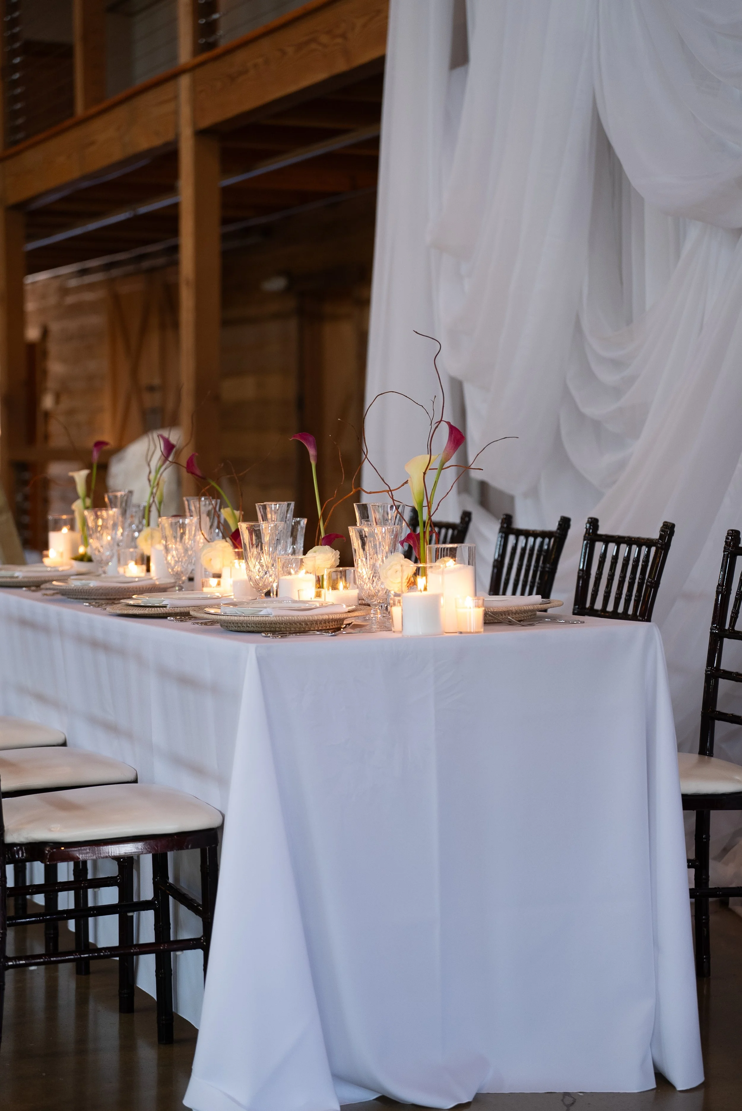 A decorated banquet table with white tablecloth, floral arrangements, candles, and black chairs, set in a rustic venue with wooden walls and drapery.
