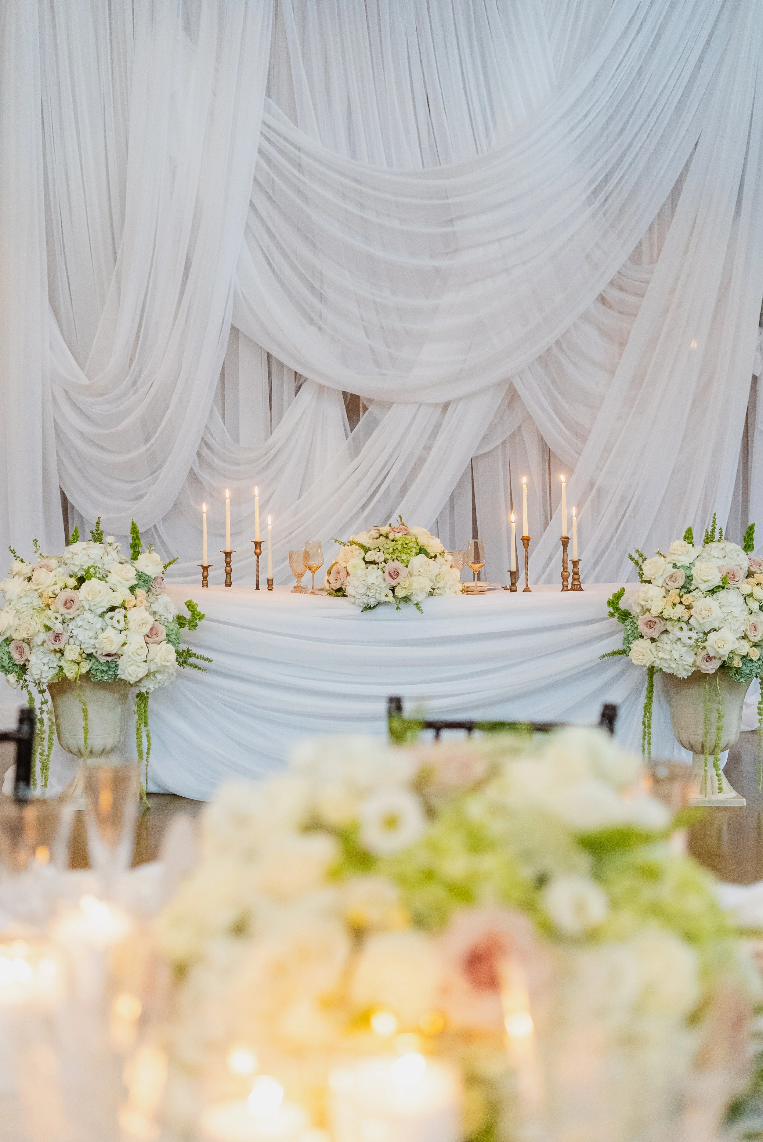 Elegant wedding reception table with white draped fabric backdrop, floral arrangements, and lit candles.