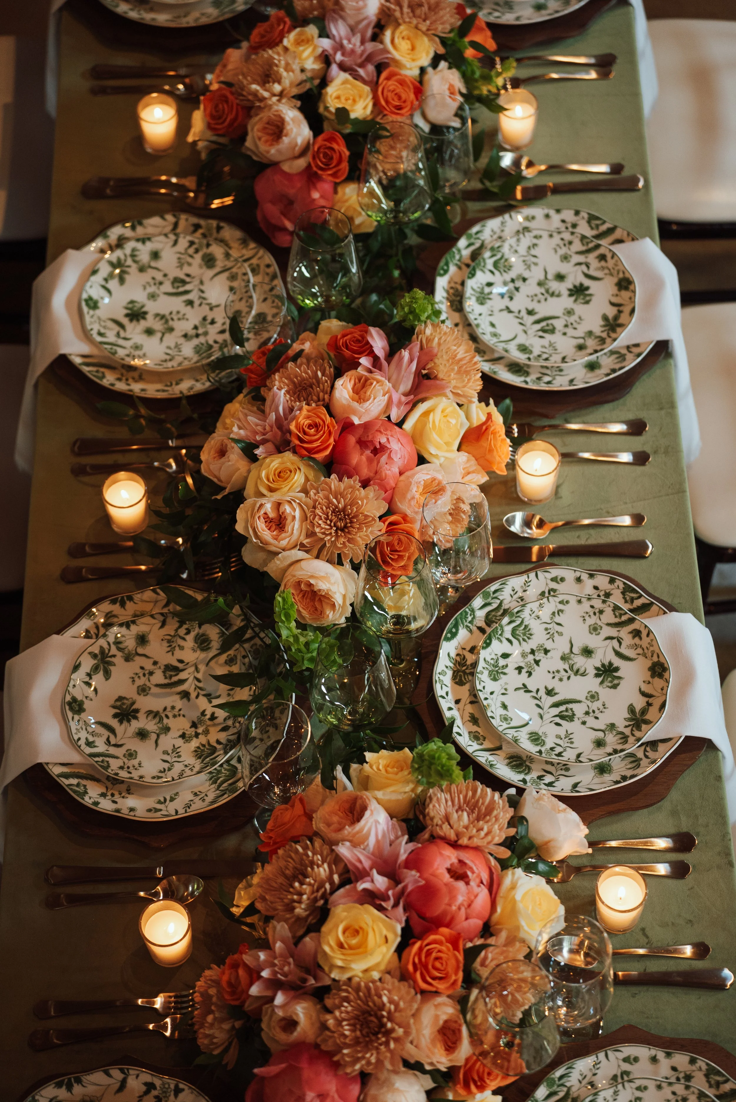 Elegant dining table with floral centerpiece, candles, and herbal-patterned plates arranged for a celebration.
