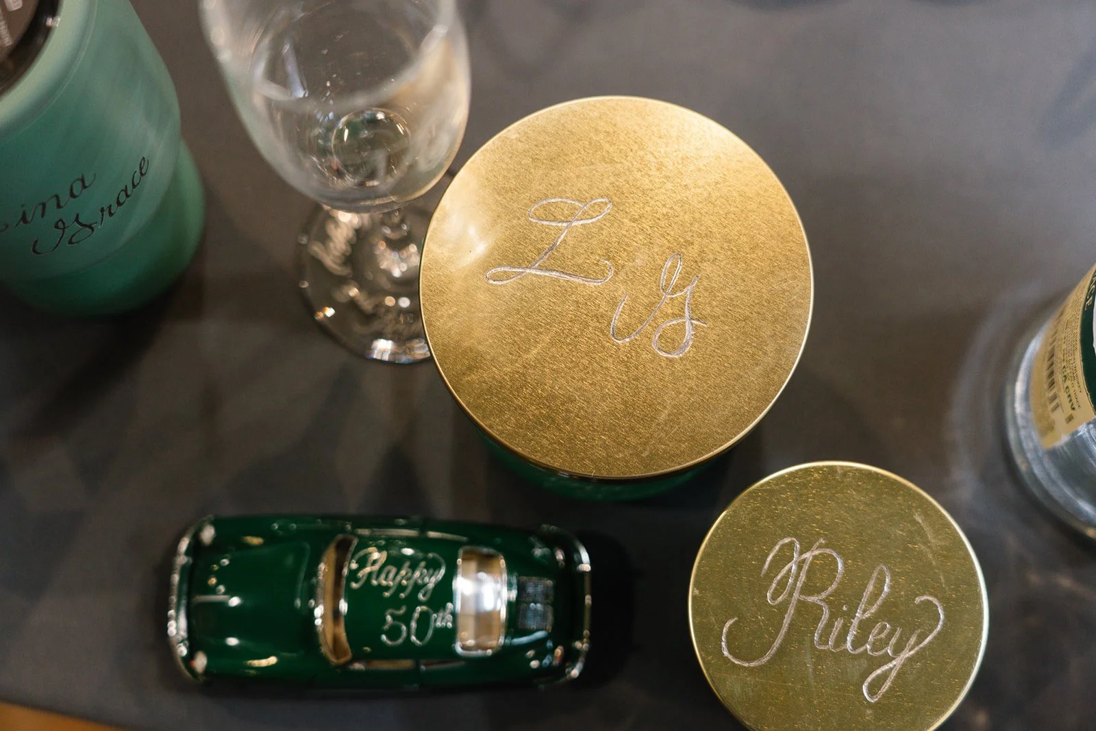 Decorative celebration items including a small green toy car with a gold label, two gold-edged container lids with names "LJS" and "Riley" written on them, a partially visible party bottle, a glass of water, and a turquoise container with 