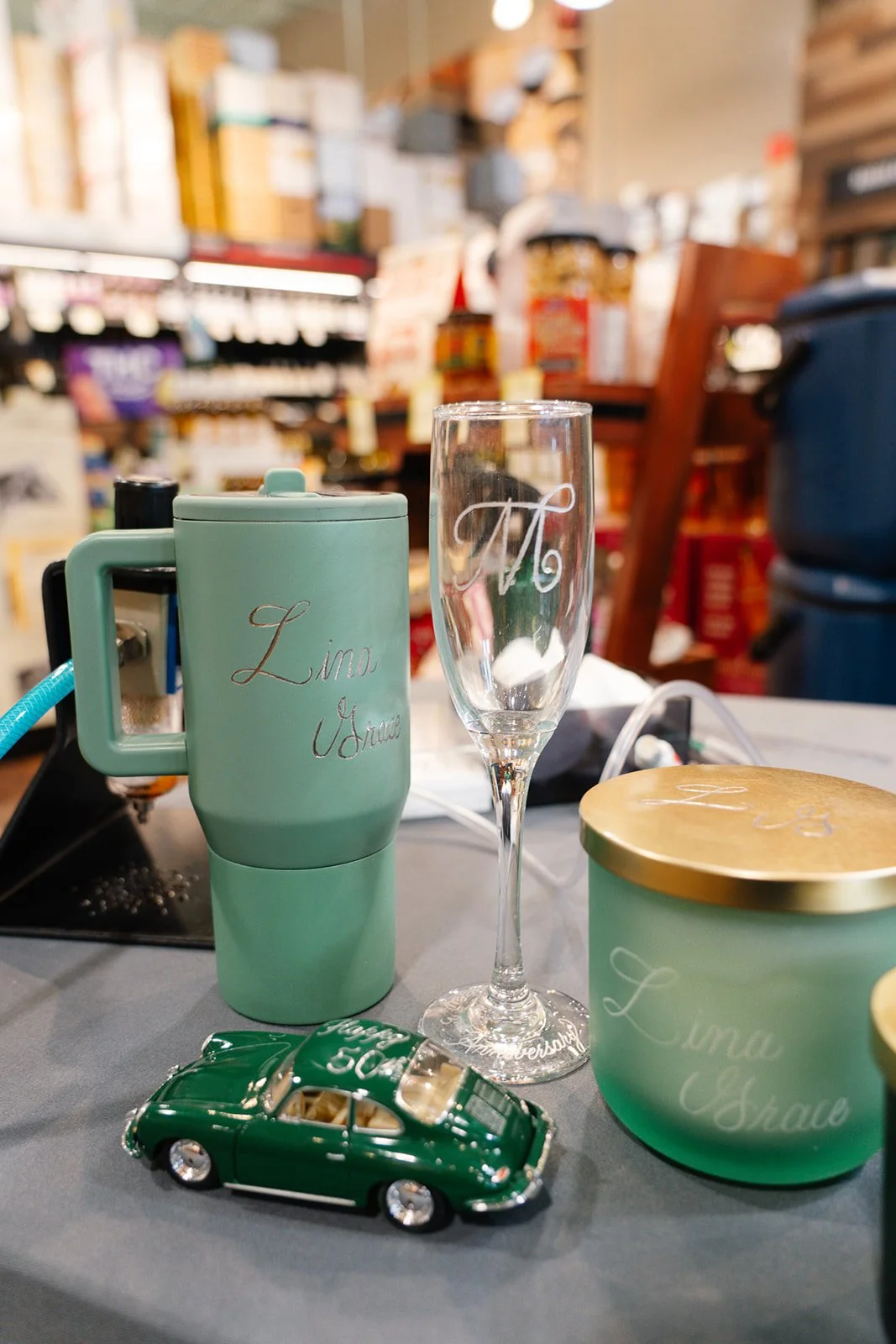 A green insulated mug with hand engraving calligraphy on it, a clear wine glass with 'm' engraved, a green container with a gold lid with hand engraving name