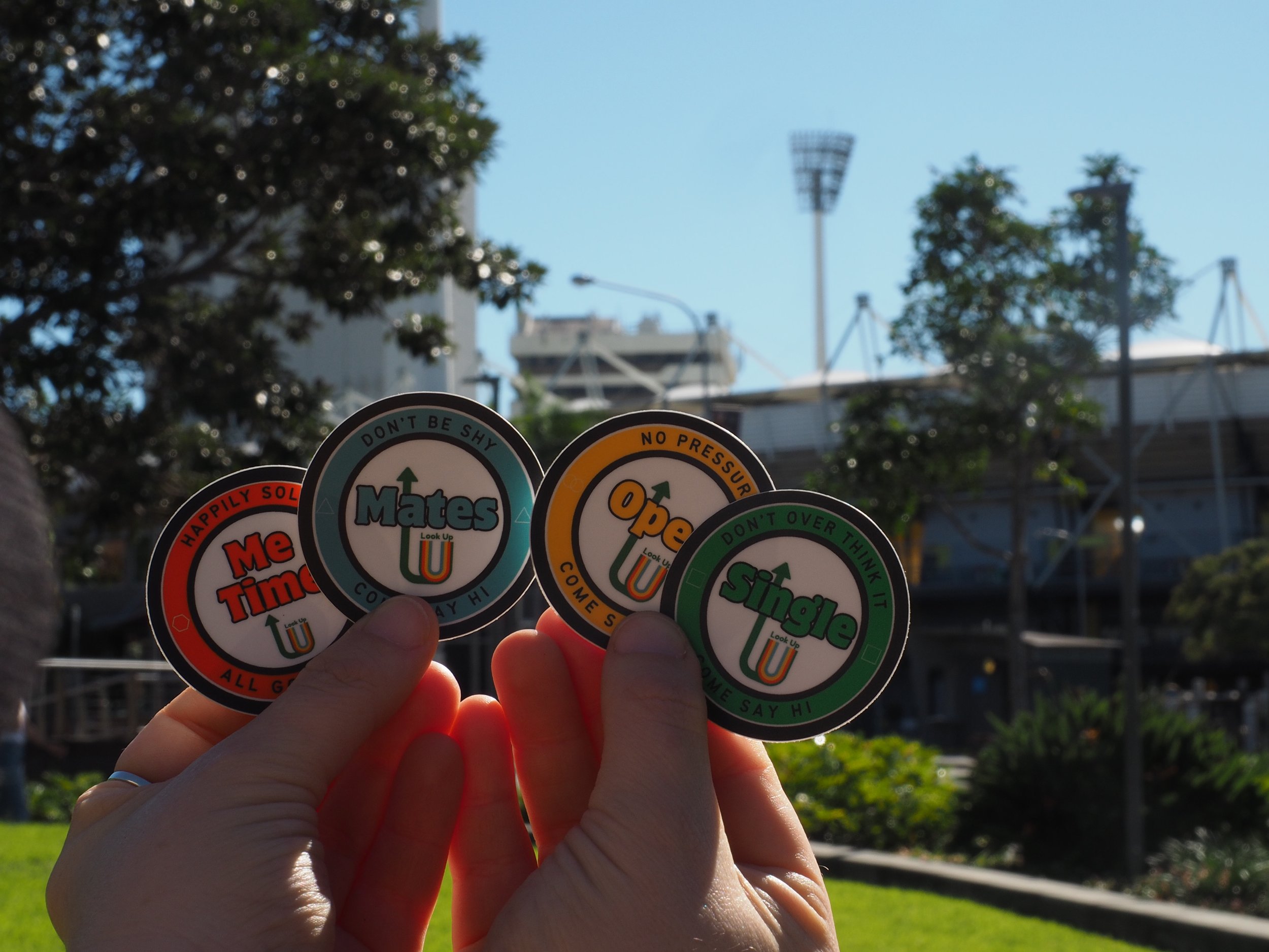 A person's hand holding four colorful circular stickers with motivational messages, with trees and stadium lighting in the background.